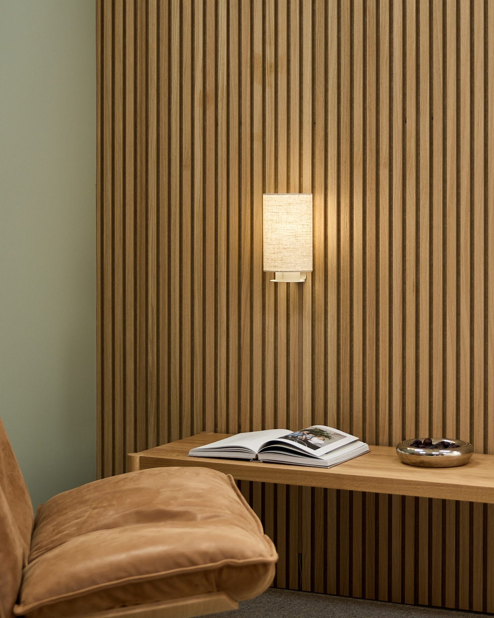 Pièce nordique minimaliste avec une chaise fauve, un mur acoustique à lames Akupanel WoodUpp, la lampe murale Feya en Beige/Beige, un livre ouvert sur un banc et un bol.