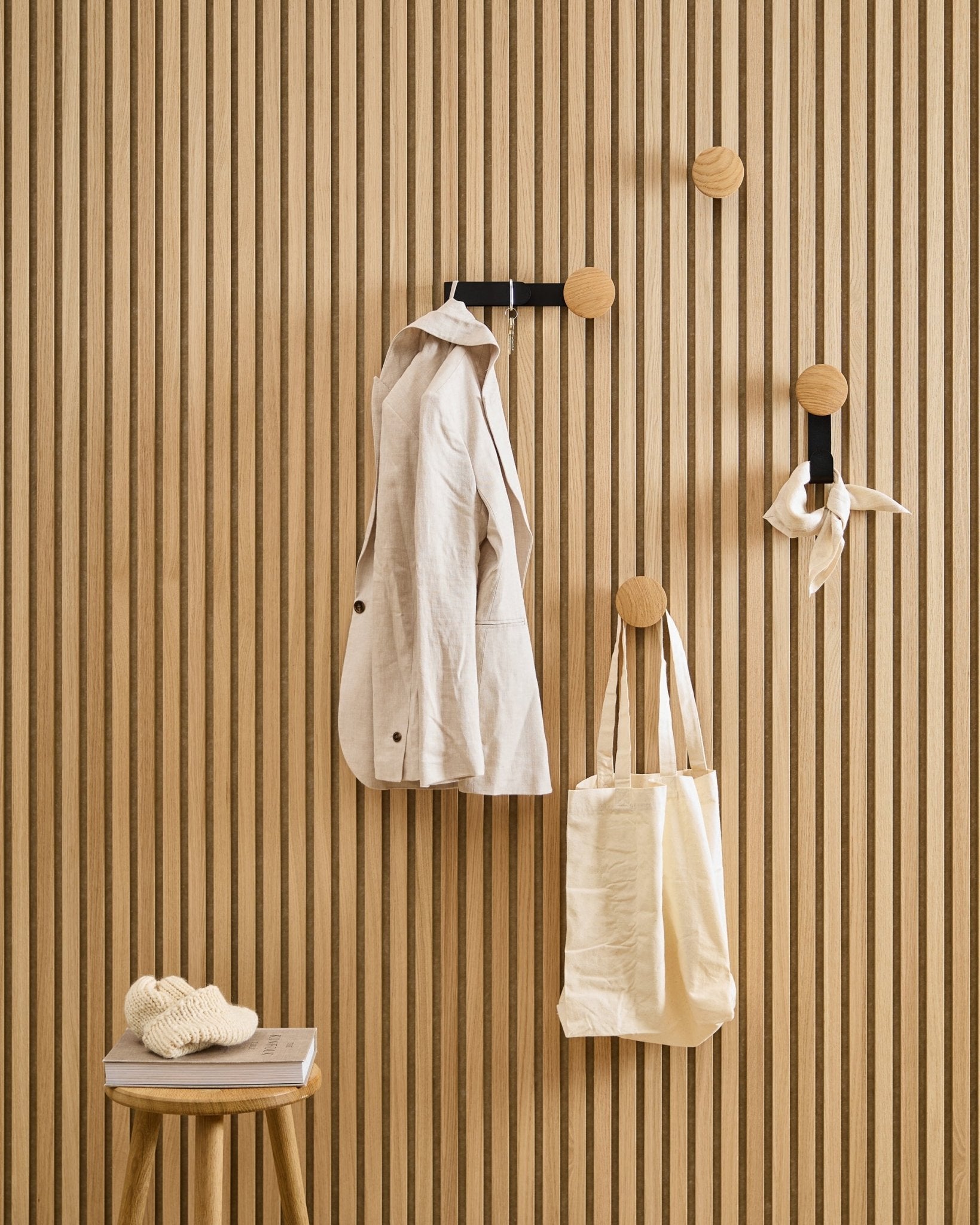 Eine Jacke in beige, eine Tragetasche und ein Schal hängen an WoodUpps Linn Horizontal Hanger in Natural Oak; auf einem Hocker daneben liegen ein Buch und ein gestricktes Teil.