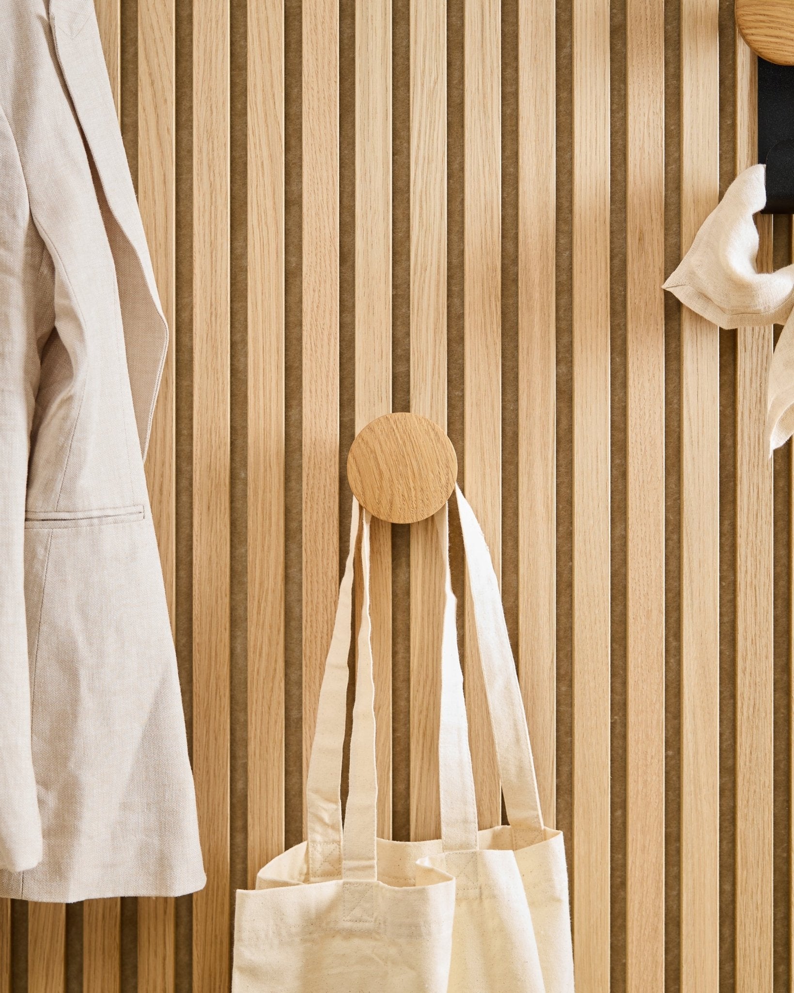 Ein beigefarbener Mantel und eine Tragetasche hängen am Linn I Single Hanger in Natural Oak von WoodUpp, montiert auf einem verticalen Akupanel Akustikpaneel aus Holzlamellen.