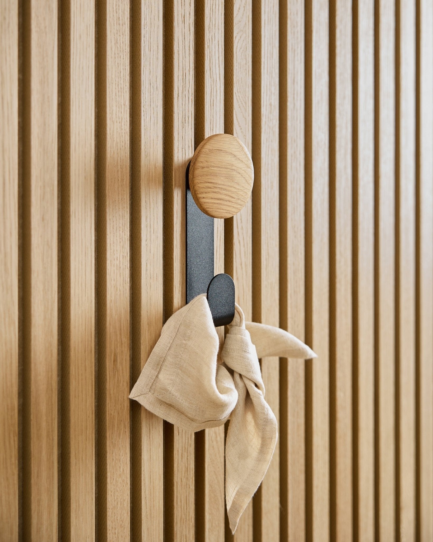 Ein beigefarbenes Tuch hängt am Linn Vertical Hanger in Natural Oak von WoodUpp, montiert an einem verticalen Akupanel Akustik-Lamellenpaneel.