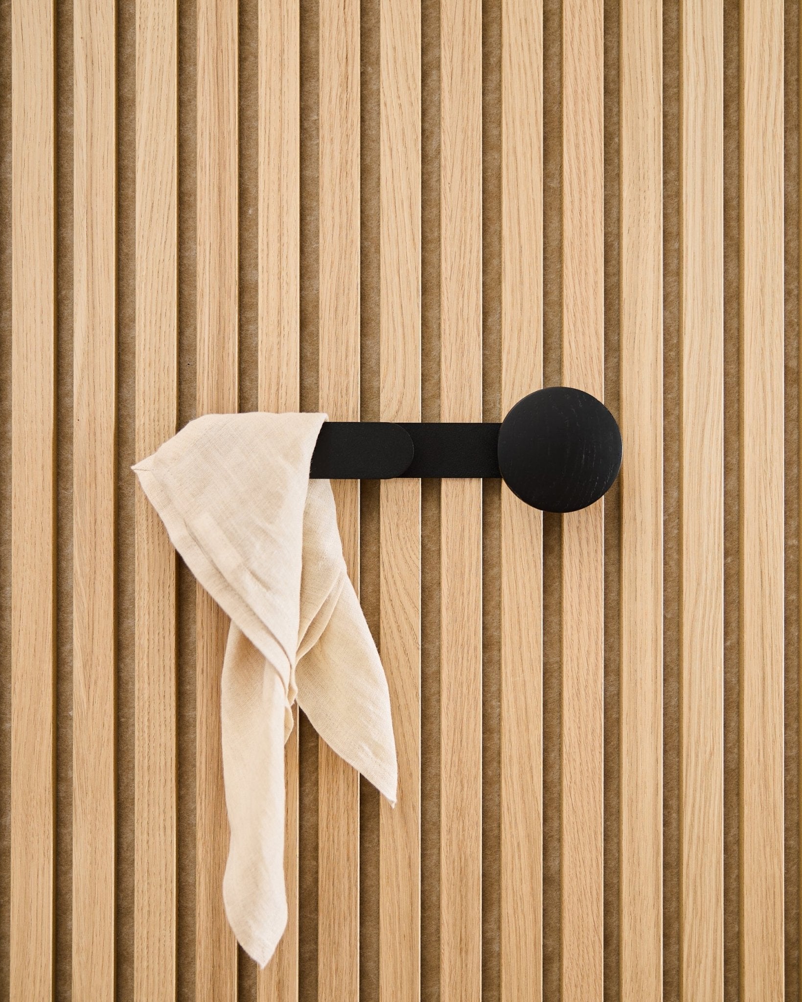 Ein beigees Tuch liegt über dem WoodUpp Linn Horizontal Hanger in Black Oak, befestigt an einem verticalen Akustikpaneel mit Holzlamellen.