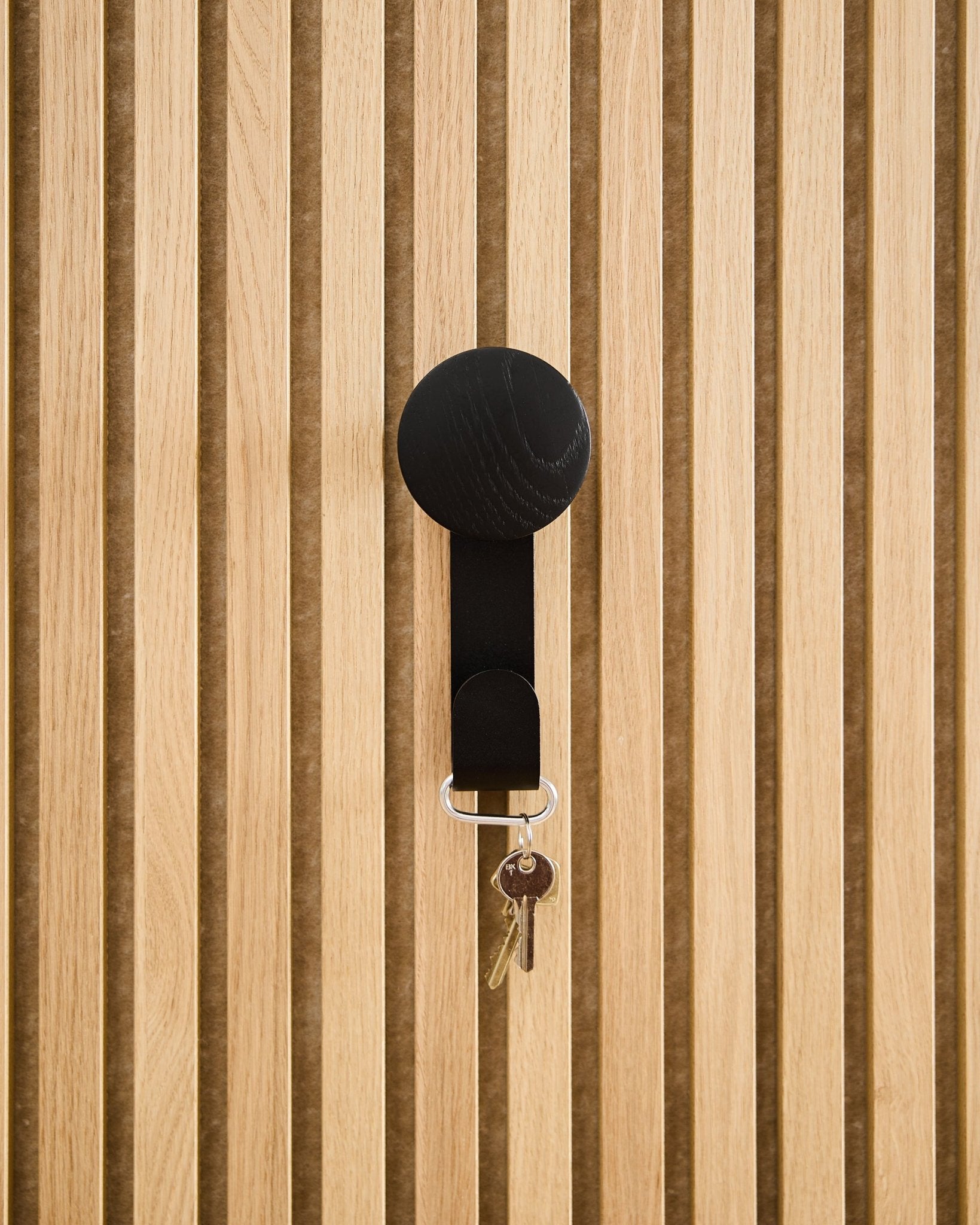 Ein Linn Vertical Hanger in Black Oak von WoodUpp trägt einen Schlüsselbund mit zwei Schlüsseln und einem runden Anhänger und ist an einem strukturierten Akupanel Akustikholzpaneel montiert.