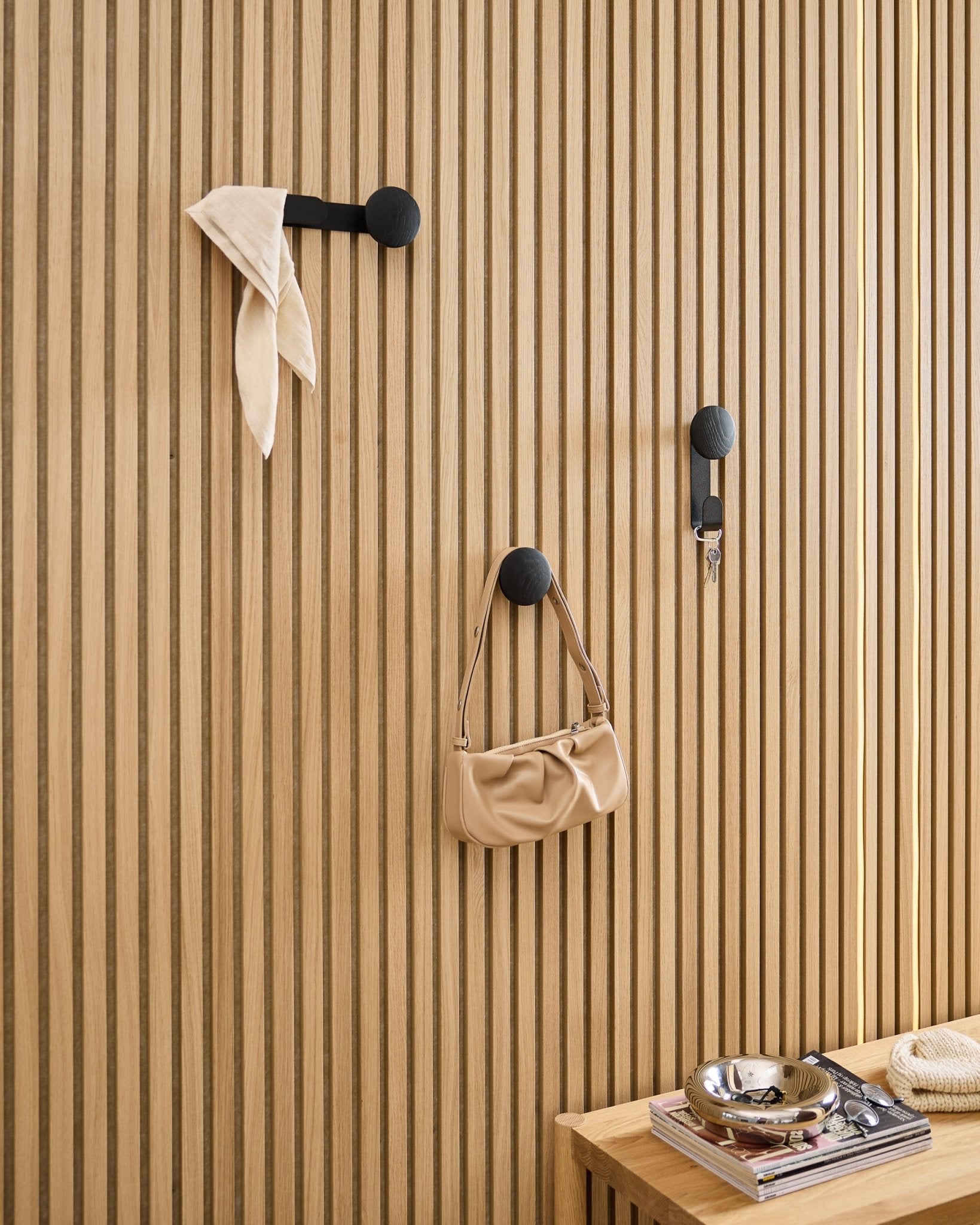 Der WoodUpp Linn Single Hanger in Black Oak trägt einen Schal, eine beige Tasche und Schlüssel an einem black Akupanel Akustik-Holzpaneel; daneben eine Bank mit Büchern und einer Metallschale.