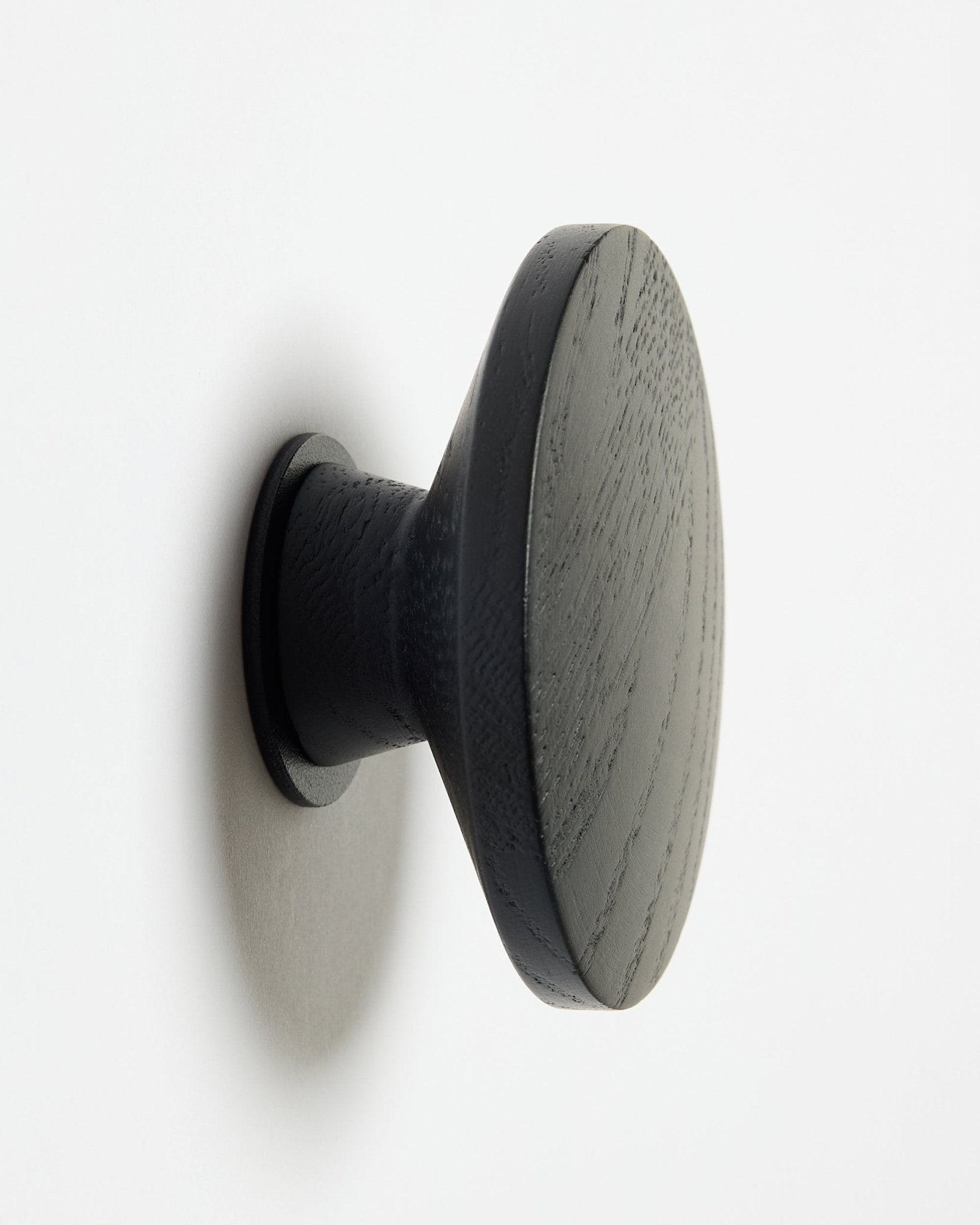 Ein runder black oak Linn | Single Hanger von WoodUpp, entworfen als Accessoire für Akupanel Akustik-Holzpaneele, montiert auf einer weißen Oberfläche mit einem weichen Schatten links.