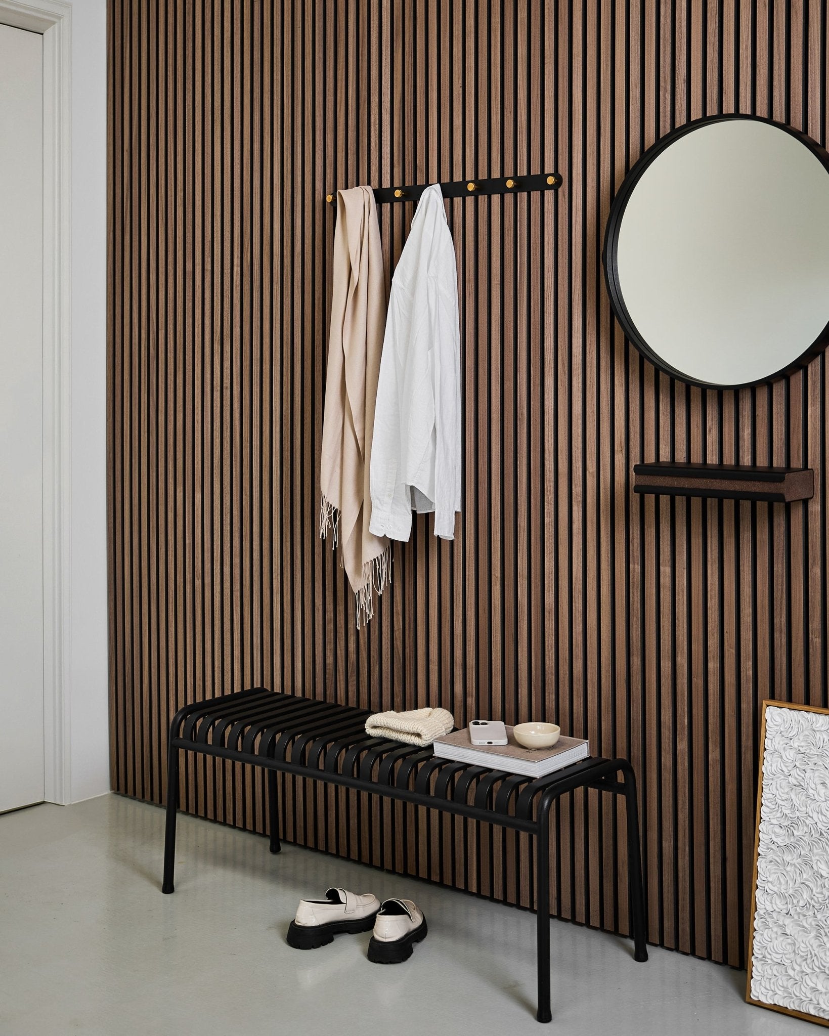 Entrée minimaliste avec un mur à lattes acoustiques WoodUpp Akupanel, le porte-manteau mural Norr (Black/Brass, X7), un banc, un miroir et une décoration moderne aux tons neutres.