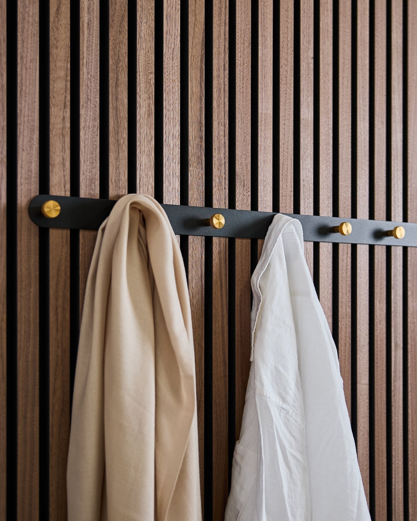 Die WoodUpp Norr Garderobenhakenleiste in black/brass (X7) mit einem beigefarbenen Schal und einem weißen Hemd hängt an einer nordischen Akustik-Lamellenwand aus Holz.