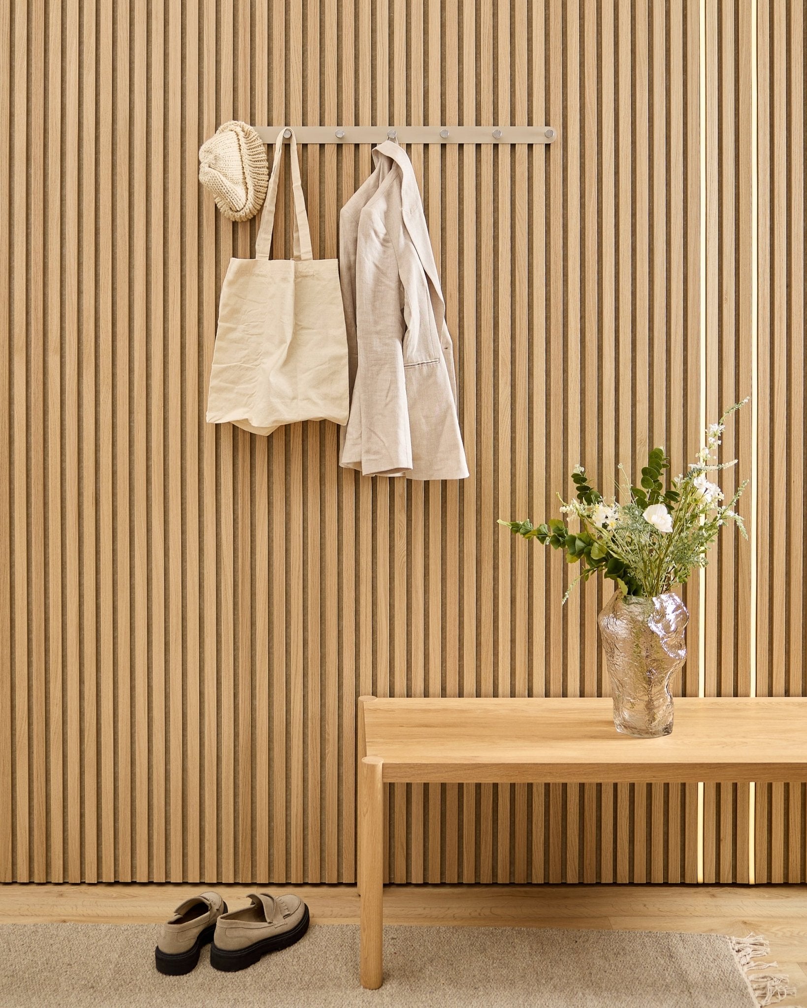 Eine beige Tasche, ein Hut und ein Hemd hängen an der WoodUpp Norr Garderobenhakenleiste über Schuhen; daneben stehen eine Bank und eine Vase neben der gestreiften Akustik-Lamellenwand.