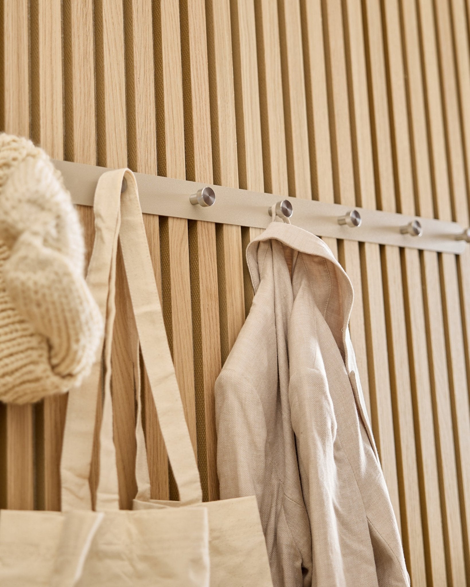 Eine beige Tragetasche, eine Strickmütze und ein Leinenhemd hängen an der WoodUpp Norr Garderobenhakenleiste (Beige/Edelstahl, X7), montiert an einer hellen Holz-Akupanel Lamellenwand.