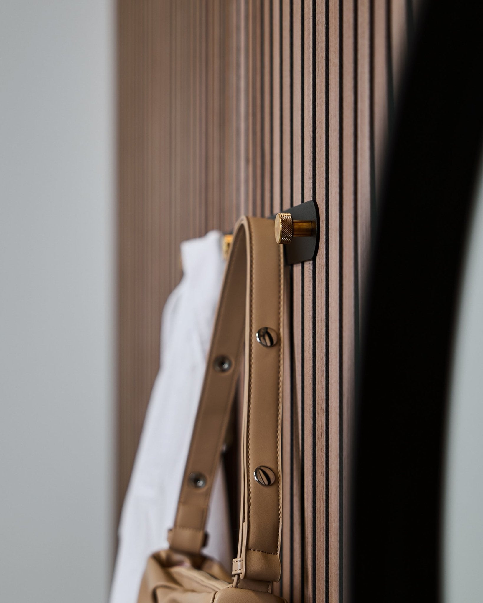Eine hellbraune Tasche und ein weißes Hemd hängen an einer WoodUpp Norr Garderobenhakenleiste (Black/Brass, X5), die an einem verticalen Akupanel Akustikpaneel mit Lamellen montiert ist.