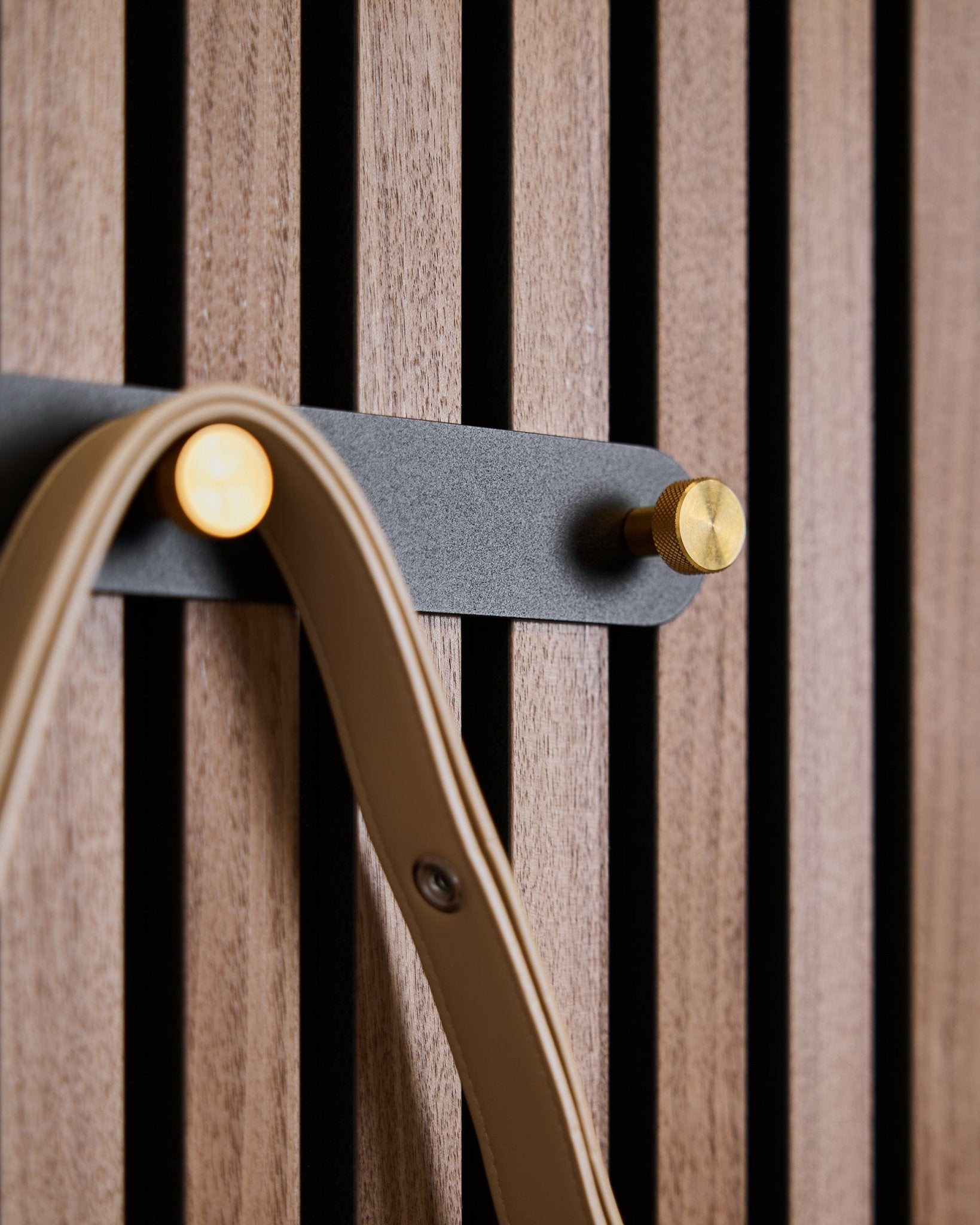 Fünf black/brass Haken der Norr Garderobenhakenleiste von WoodUpp sind auf einem Akustikpaneel aus Holz mit Lamellen montiert und halten jeweils eine beige Schlaufe.