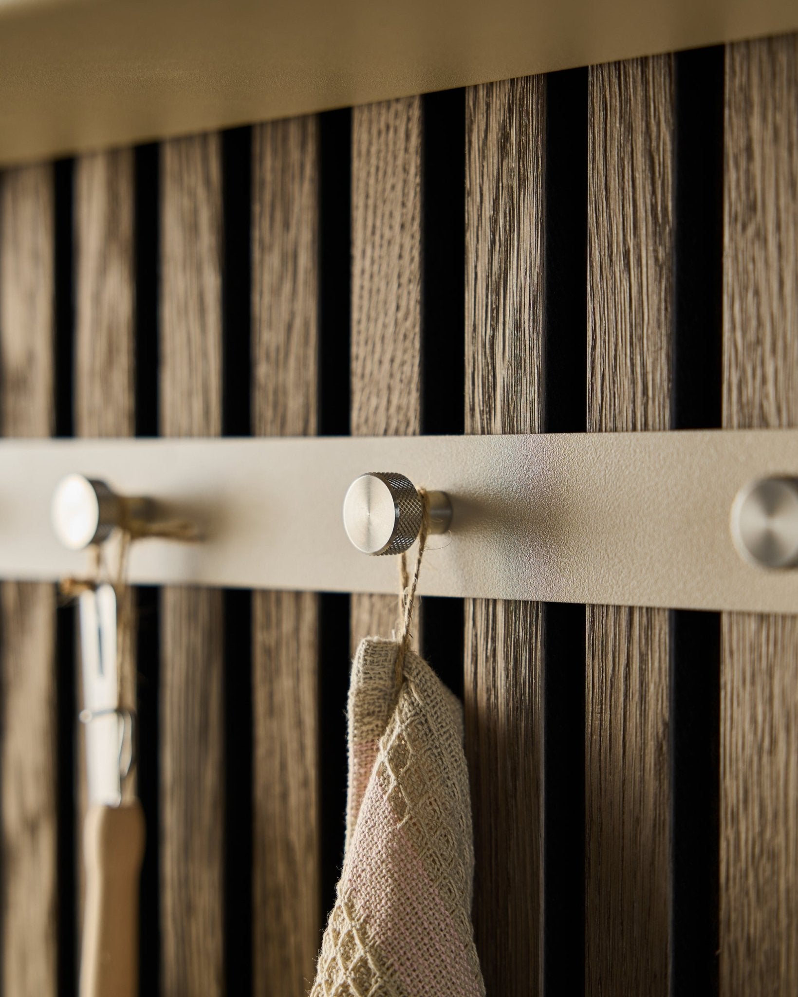 Nahaufnahme des WoodUpp Norr Coat Hook Rack (Beige/rostfreier Steel, X5), montiert auf einem Akustikpaneel mit Holzlamellen, an dem ein Handtuch und ein Werkzeug mit Griff hängen.