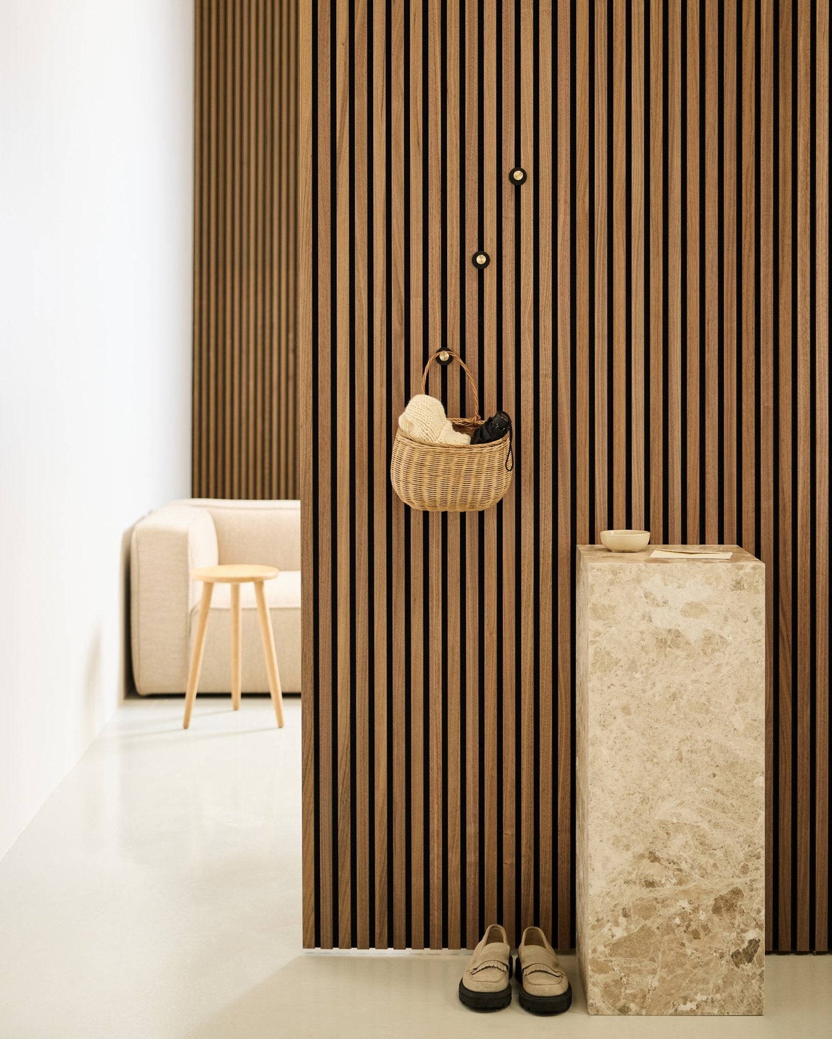 Entrée minimaliste avec mur à lattes acoustique Akupanel, patère WoodUpp Norr en black et brass, panier, piédestal en pierre, chaussons et chaise beige.