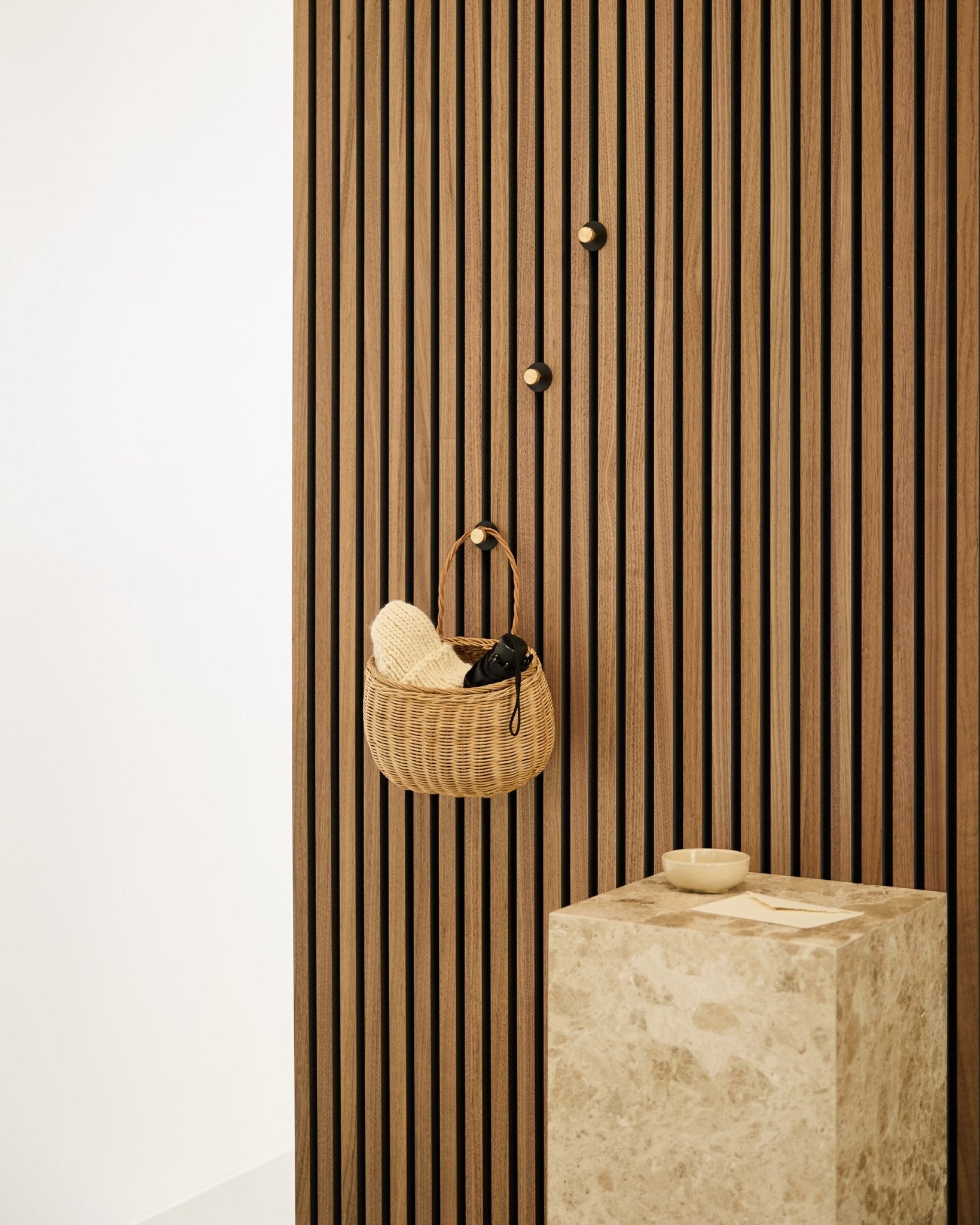 Norr Garderobenhaken von WoodUpp, montiert auf einer nordischen Akupanel Akustik-Lamellenwand, neben einem beigefarbenen Steinsockel mit einer smallen Schale in einem minimalistischen Interieur.