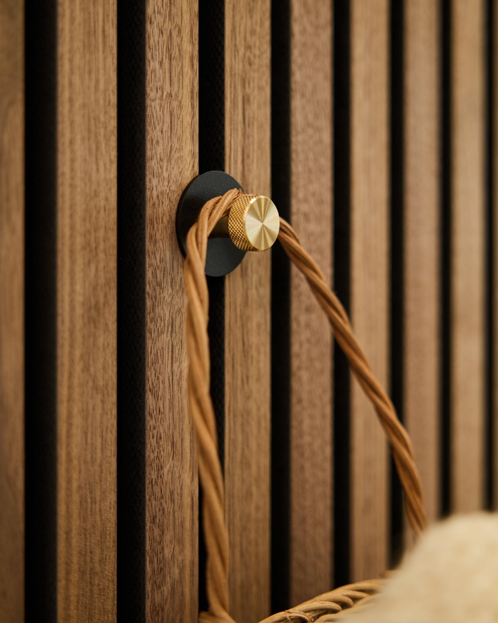 WoodUpp Norr Garderobenhaken in black und brass, montiert auf einem Holz-Akupanel Akustikpaneel, hält einen hellbraunen geflochtenen Griff.