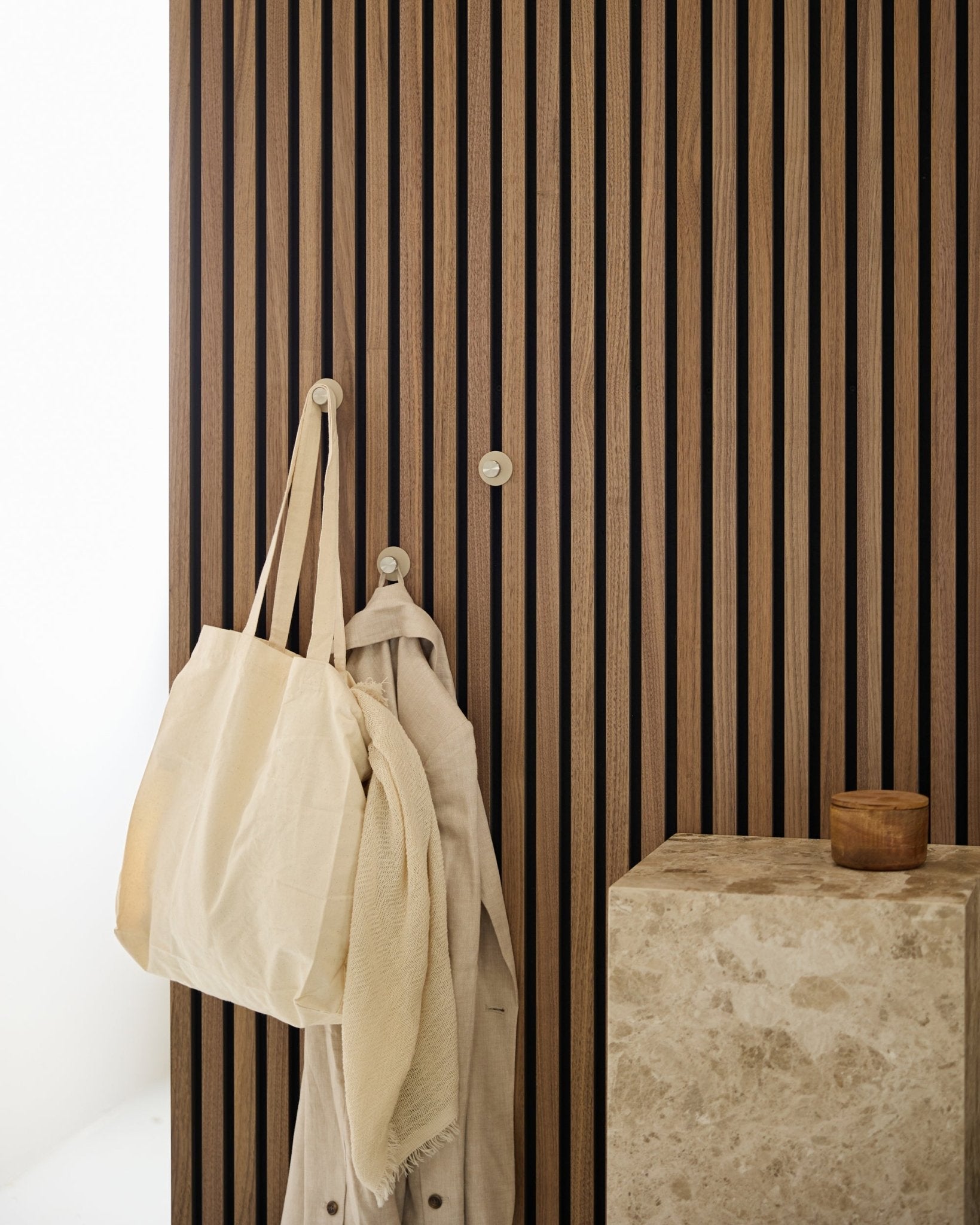 Un sac cabas beige et un tissu léger sont suspendus à une patère WoodUpp Norr (Beige/Steel inoxydable) montée sur un vertical Akupanel Panneau acoustique à côté d’une surface en pierre.