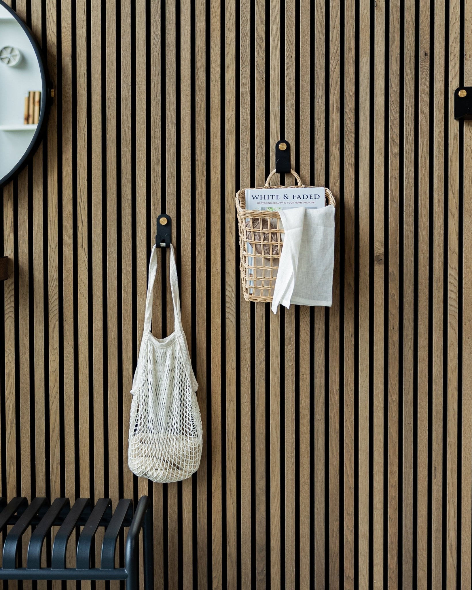 Un sac en filet et un panier sont suspendus à des Norr Single Hangers de WoodUpp sur un Akupanel en bois nordique, Panneau acoustique ; une partie d’un miroir est visible à gauche.