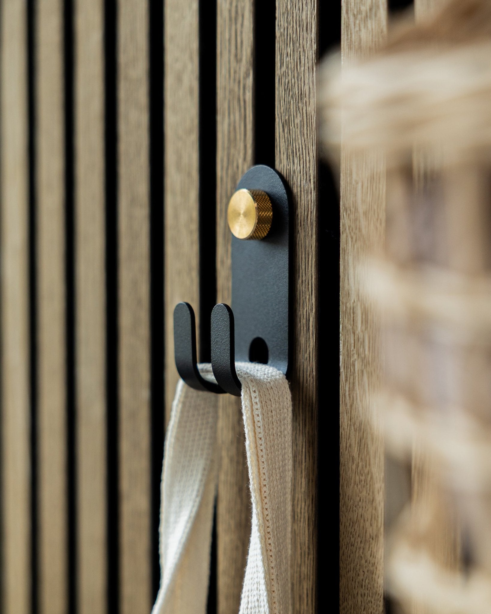 Le WoodUpp Norr Double Hanger en black est fixé sur un panneau mural acoustique à lattes en bois nordique et supporte une sangle en tissu crème-colored.