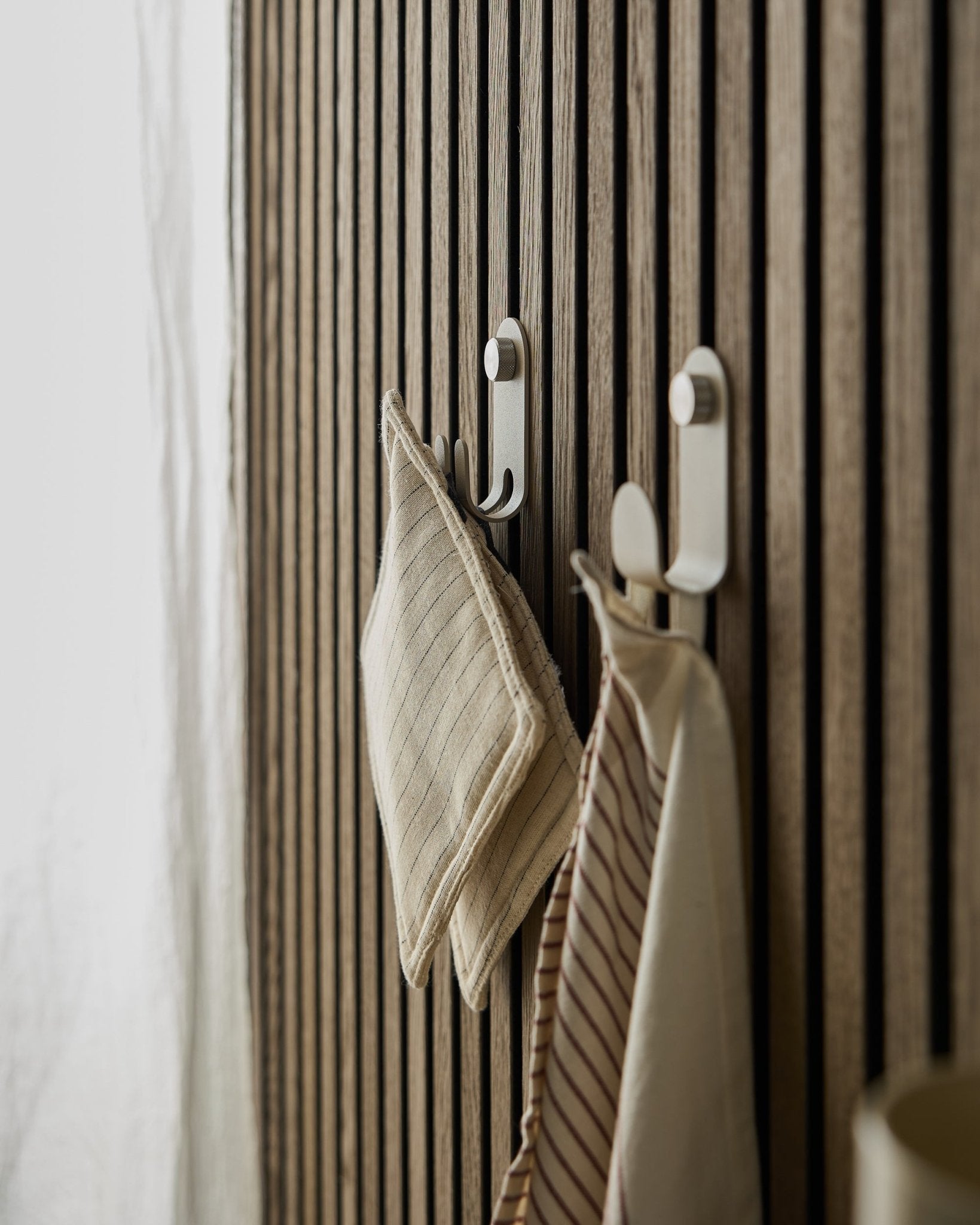 Zwei gestreifte Küchentücher hängen am Norr Double Hanger von WoodUpp, montiert auf einem nordischen Lamellenwand-Akustikpaneel in beige, beleuchtet von weichem natürlichem Licht.