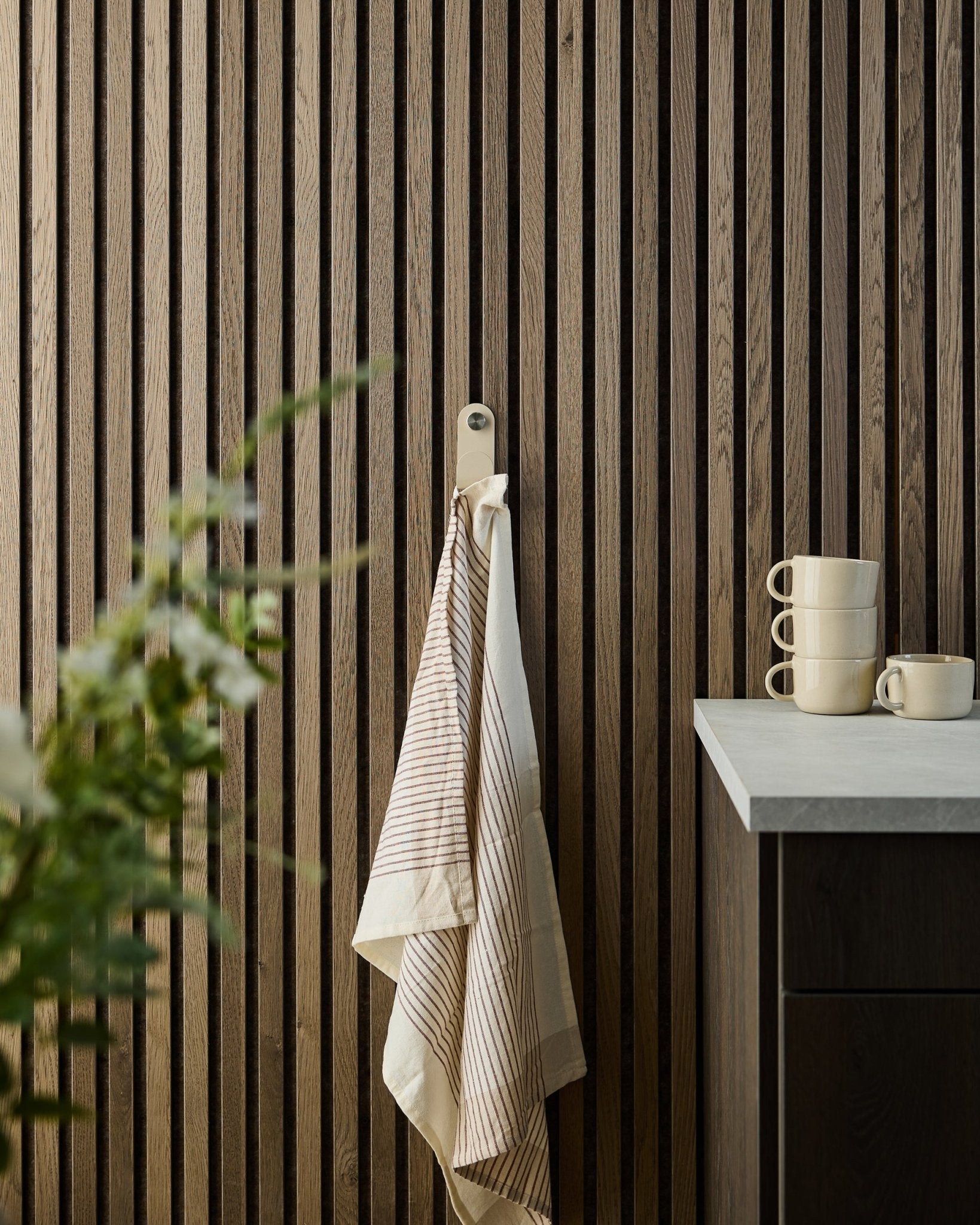 Une serviette rayée est suspendue à un WoodUpp beige Norr Single Hanger fixé sur un Panneau acoustique à lattes de bois ; des tasses empilées et une plante se trouvent sur le comptoir en dessous.
