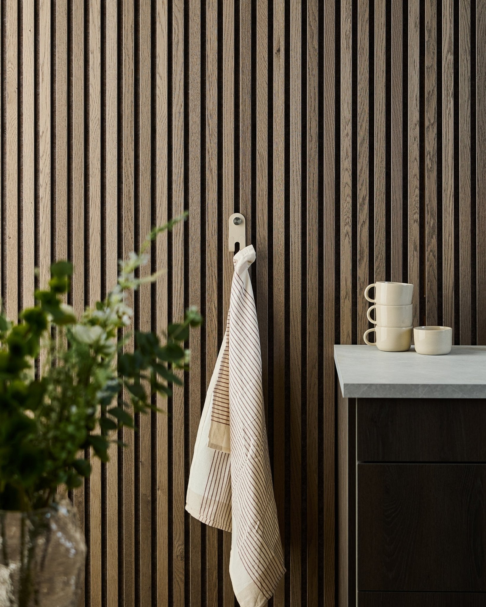 Une serviette rayée est suspendue au WoodUpp Norr Double Hanger en beige, monté sur un Panneau acoustique nordique à lattes ; des mugs, un bol et une plante verte sont posés en dessous.