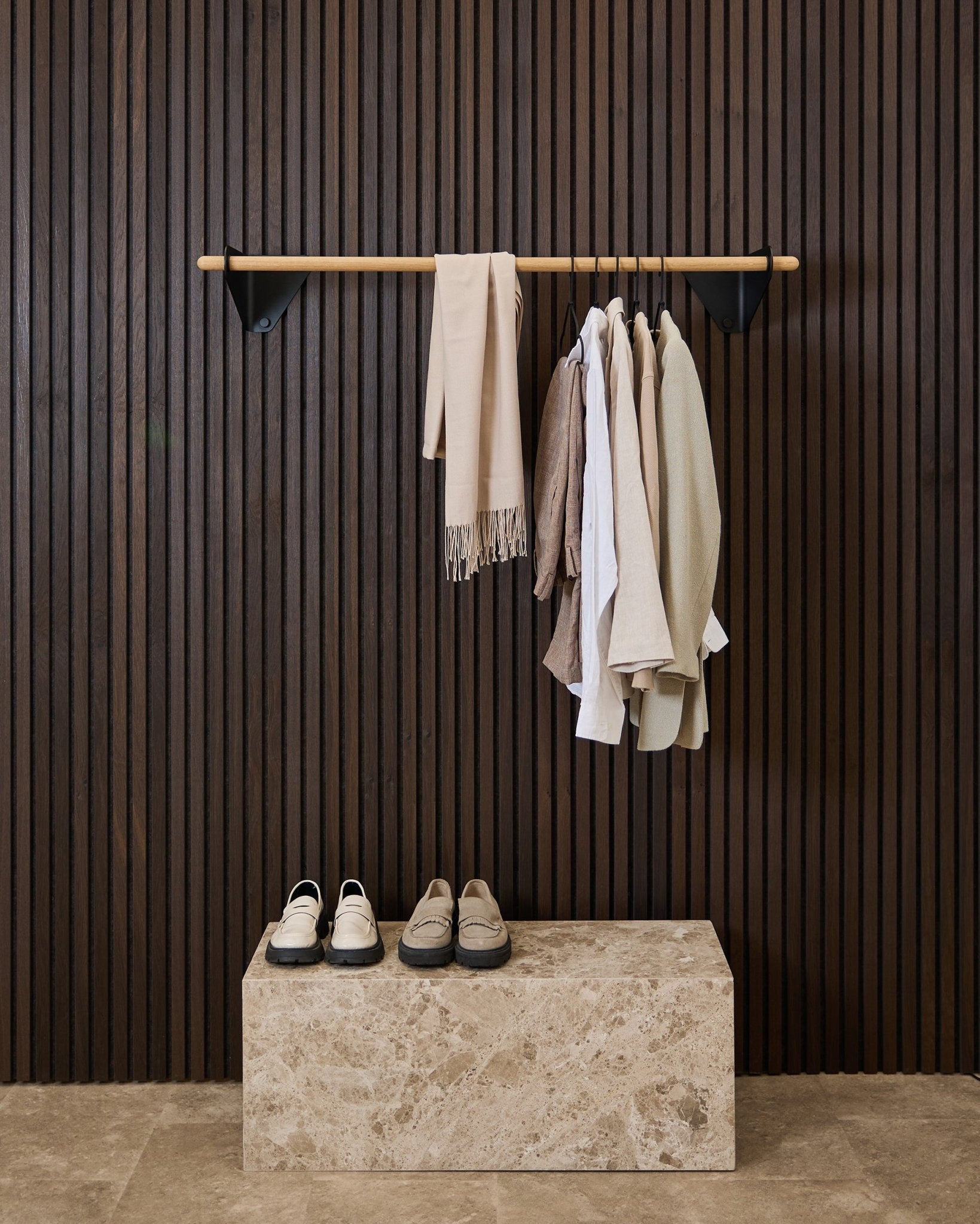 Kleidung in neutralen Tönen und ein Schal hängen an einer black 115,9 cm Linn | Coat Rail von WoodUpp über einer Marmorbank, vor einem dunklen Akupanel Akustik-Lamellenwandpaneel.