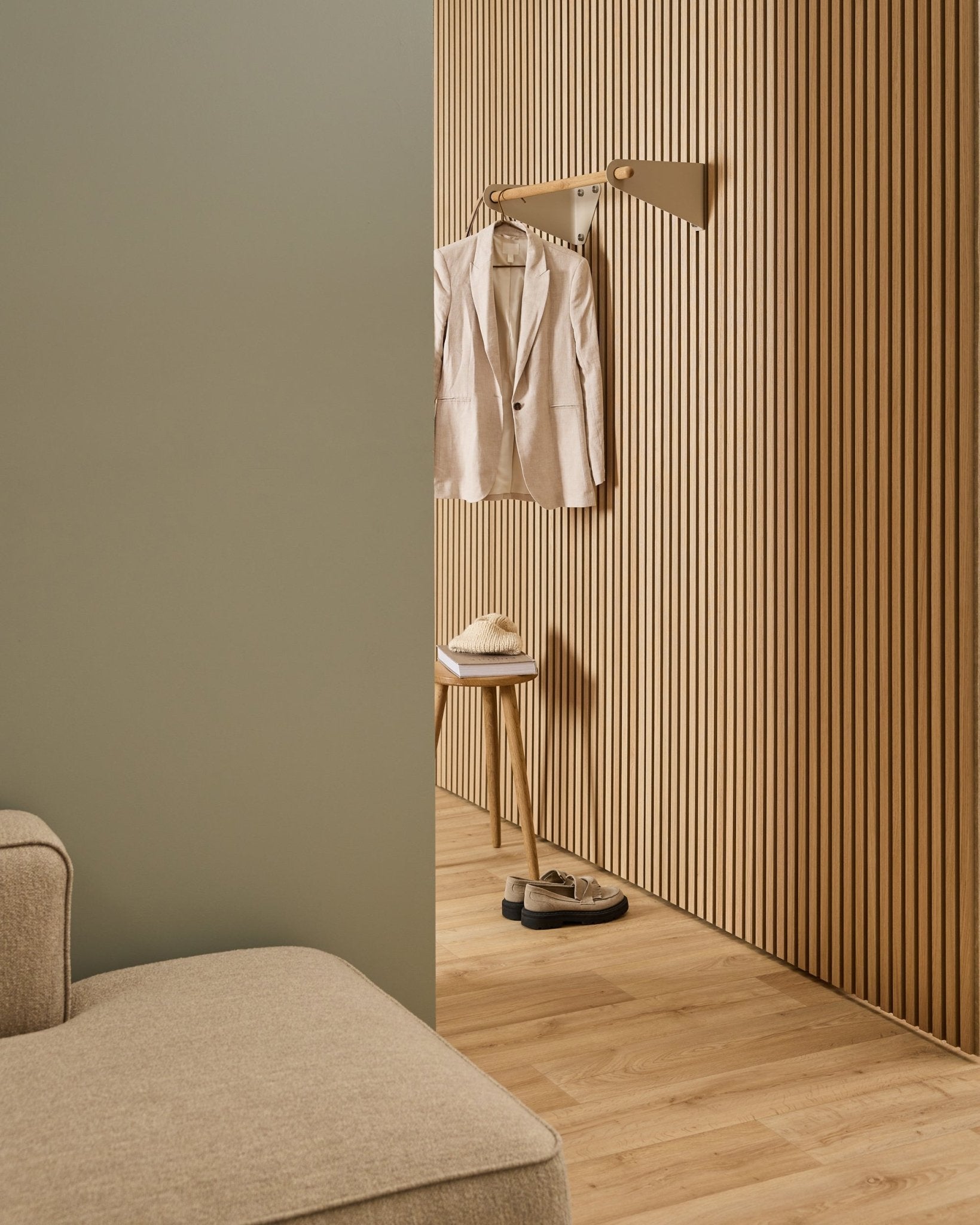 Minimalistischer Raum mit beigefarbenem Sofa, Akupanel Akustik-Lamellenwand, WoodUpp Linn Garderobenstange in beige (75,9 cm), Hocker mit Hut und Schuhen auf hellem Holzboden.