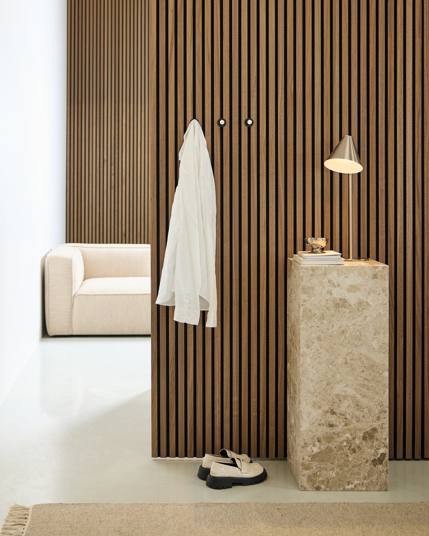 Pièce minimaliste avec un mur acoustique en tasseaux de bois Akupanel au style nordique, une chemise blanche suspendue à des crochets muraux, un piédestal en pierre avec une lampe et des livres, un canapé beige et des chaussures blanches au sol.