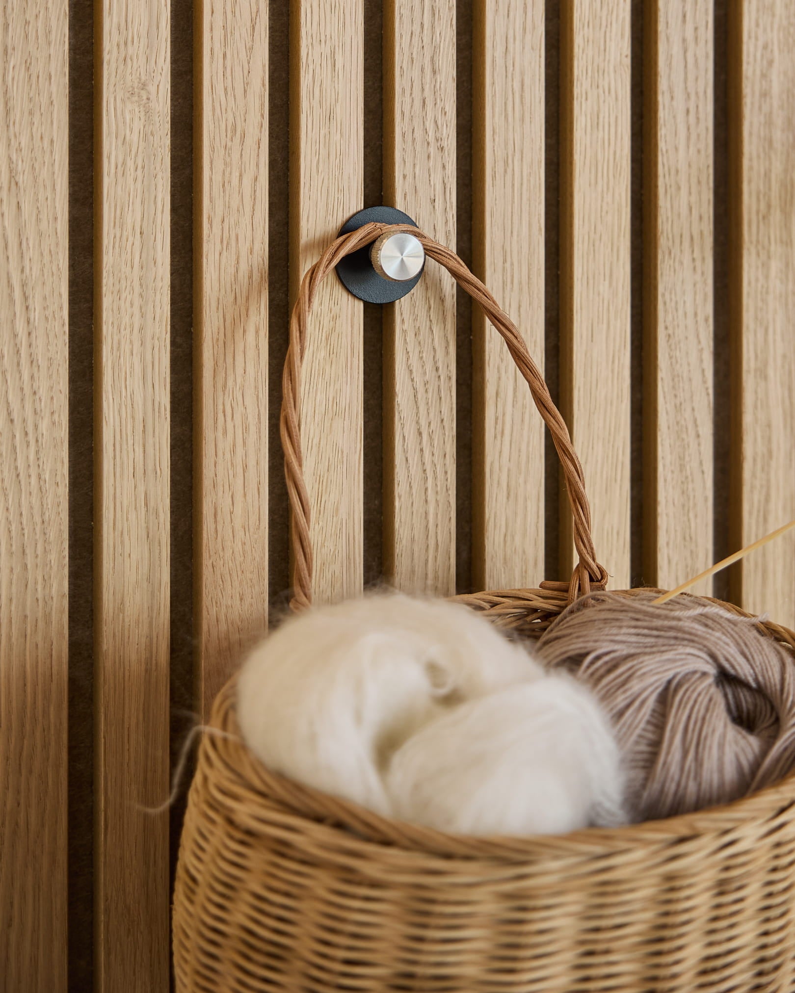 Ein geflochtener Garnkorb in Weiß und beige hängt an einem Norr Coat Hook / X1, Black/Stainless Steel von WoodUpp, montiert auf einem Akupanel Akustikpaneel mit Lamellen.