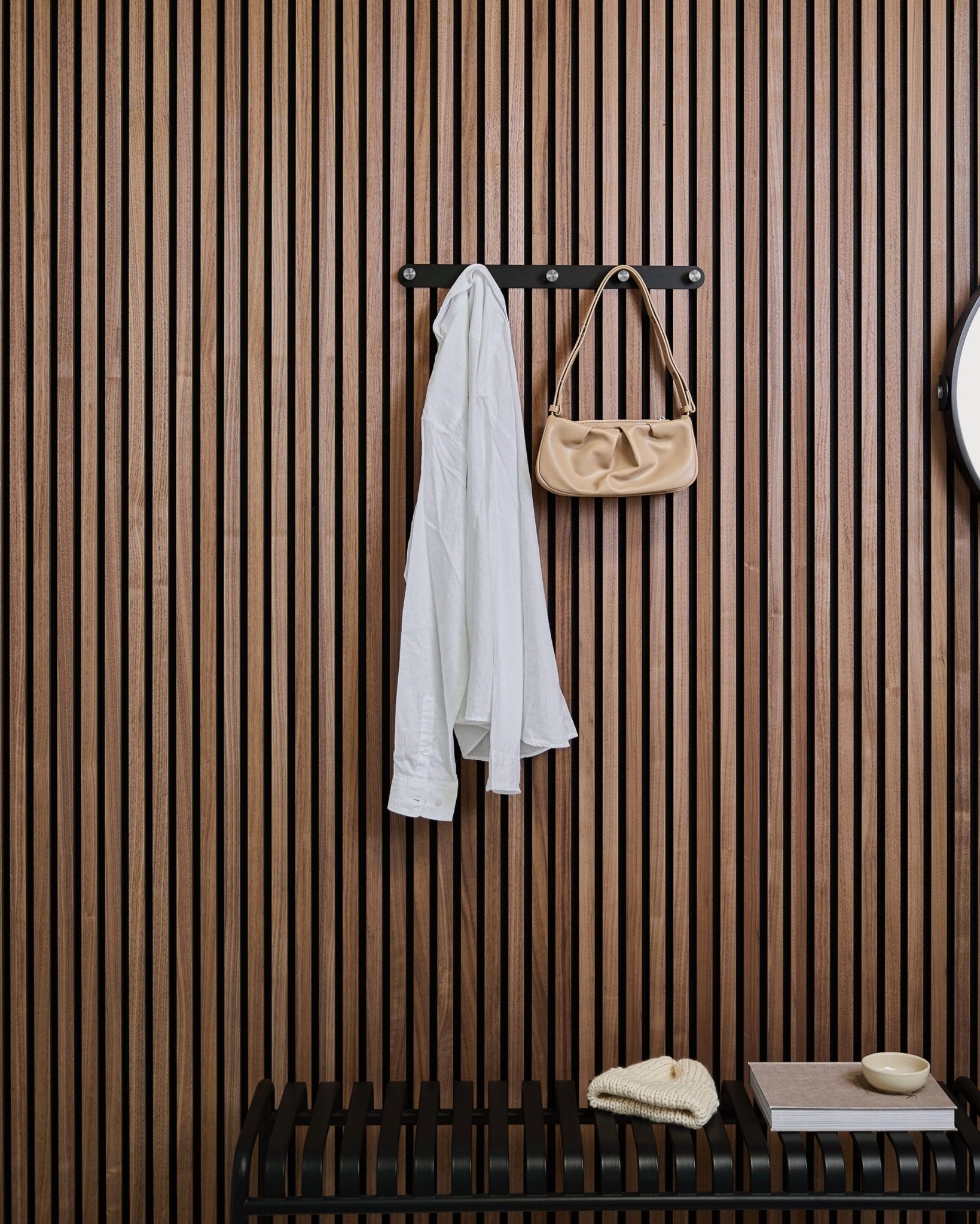 Une chemise blanche et un sac à main beige sont suspendus à la patère WoodUpps Norr au-dessus d’un banc, sur fond de panneau mural à lattes en bois Akupanel avec quelques objets déco.