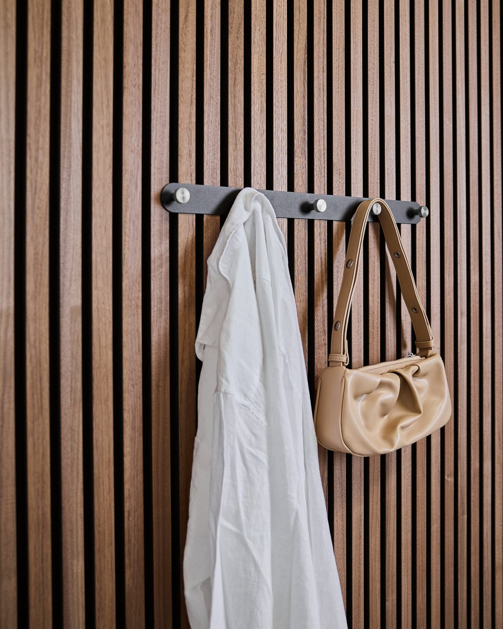 Ein weißes Hemd und eine beige Handtasche hängen an den WoodUpp Norr X5 Black/Stainless Steel Mantelhaken, die an einem vertical Akustik-Lamellenpaneel befestigt sind.