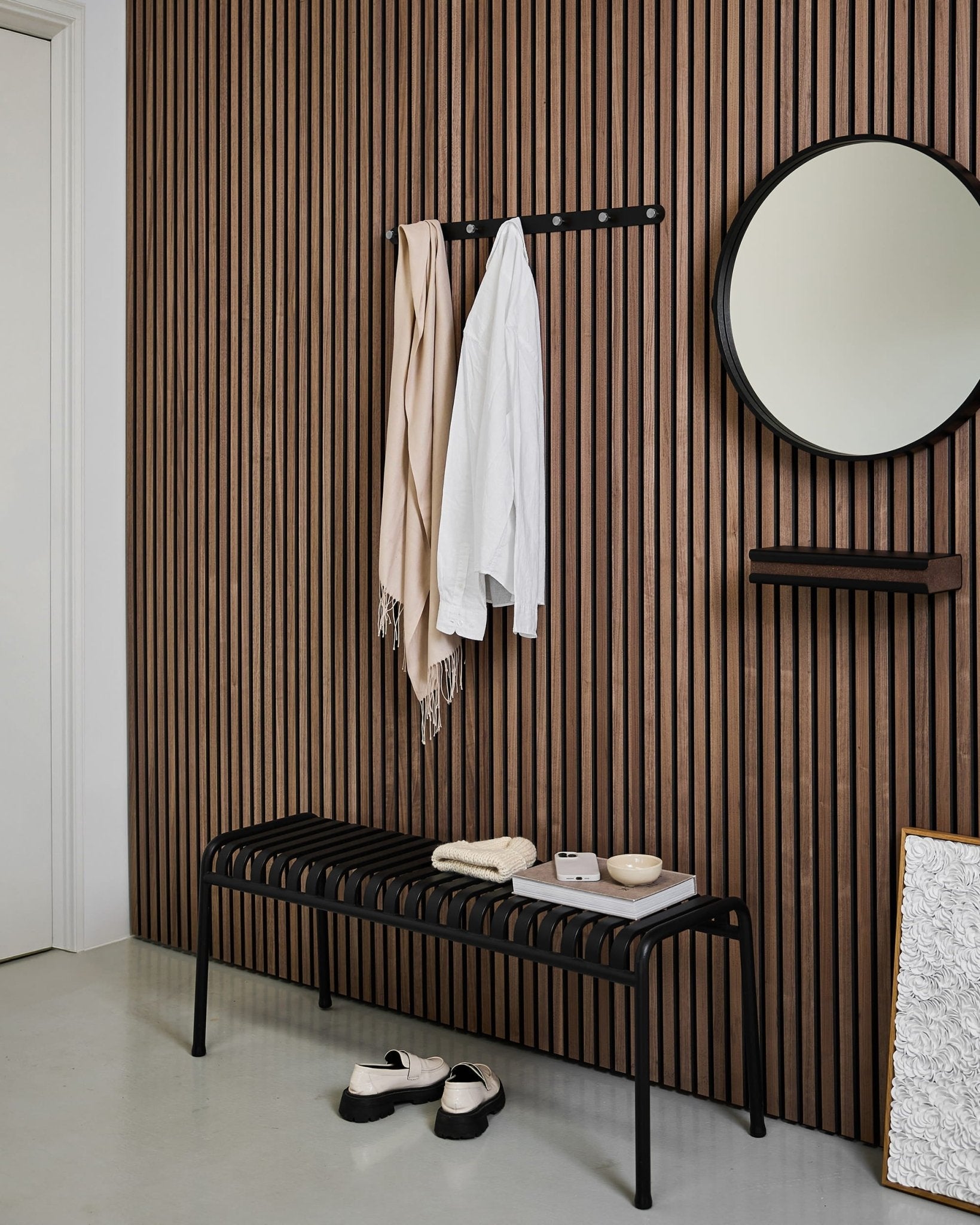 Entrée moderne avec un mur en panneau acoustique en bois à lattes de style nordique, un miroir rond, un banc black, le porte-manteau WoodUpp Norr, et des chaussons sur un sol clair.