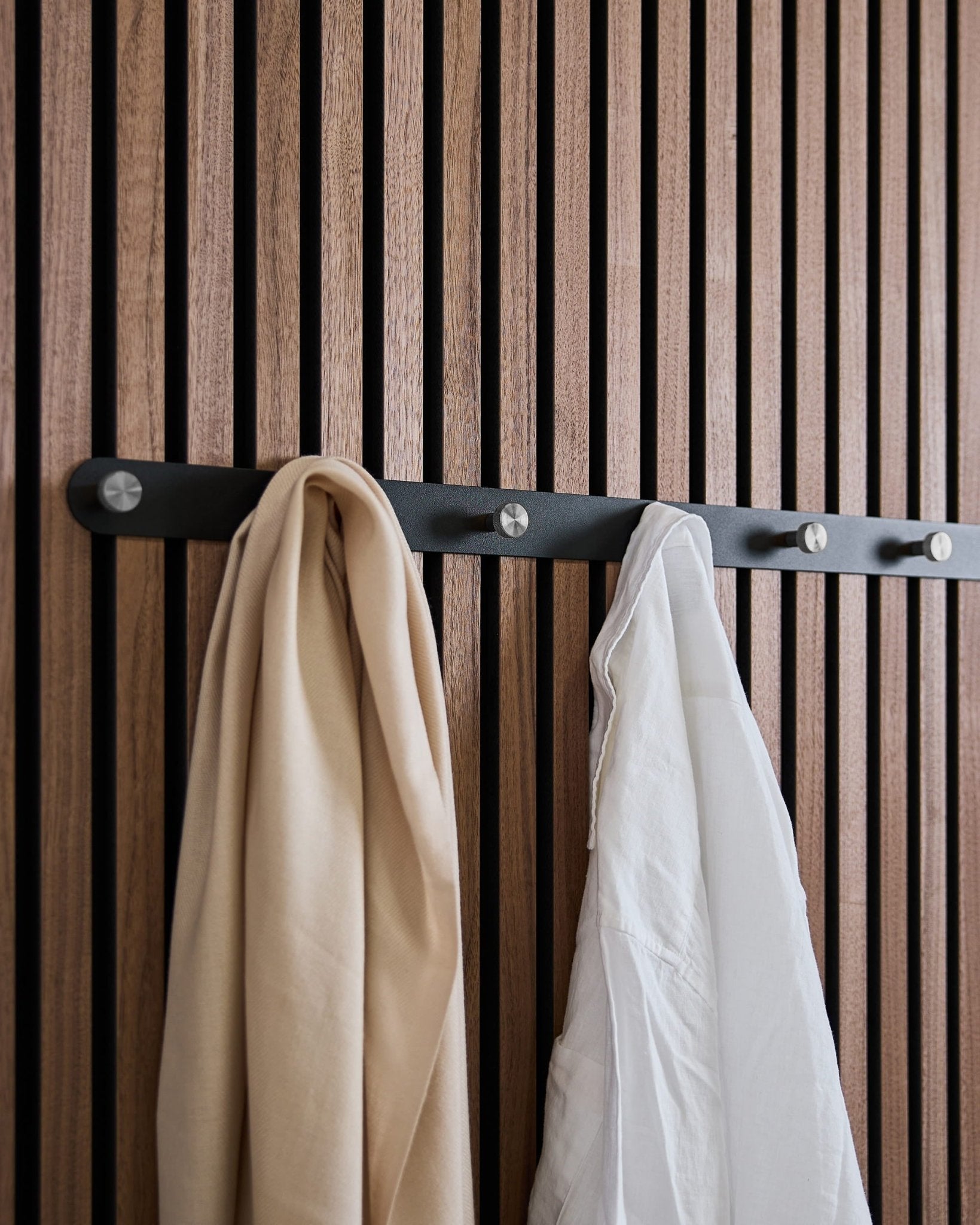 Une écharpe beige et une chemise blanche sont suspendues au porte-manteau WoodUpp Norr X7 Black/Stainless Steel, monté sur un panneau acoustique mural à lattes au style nordique.