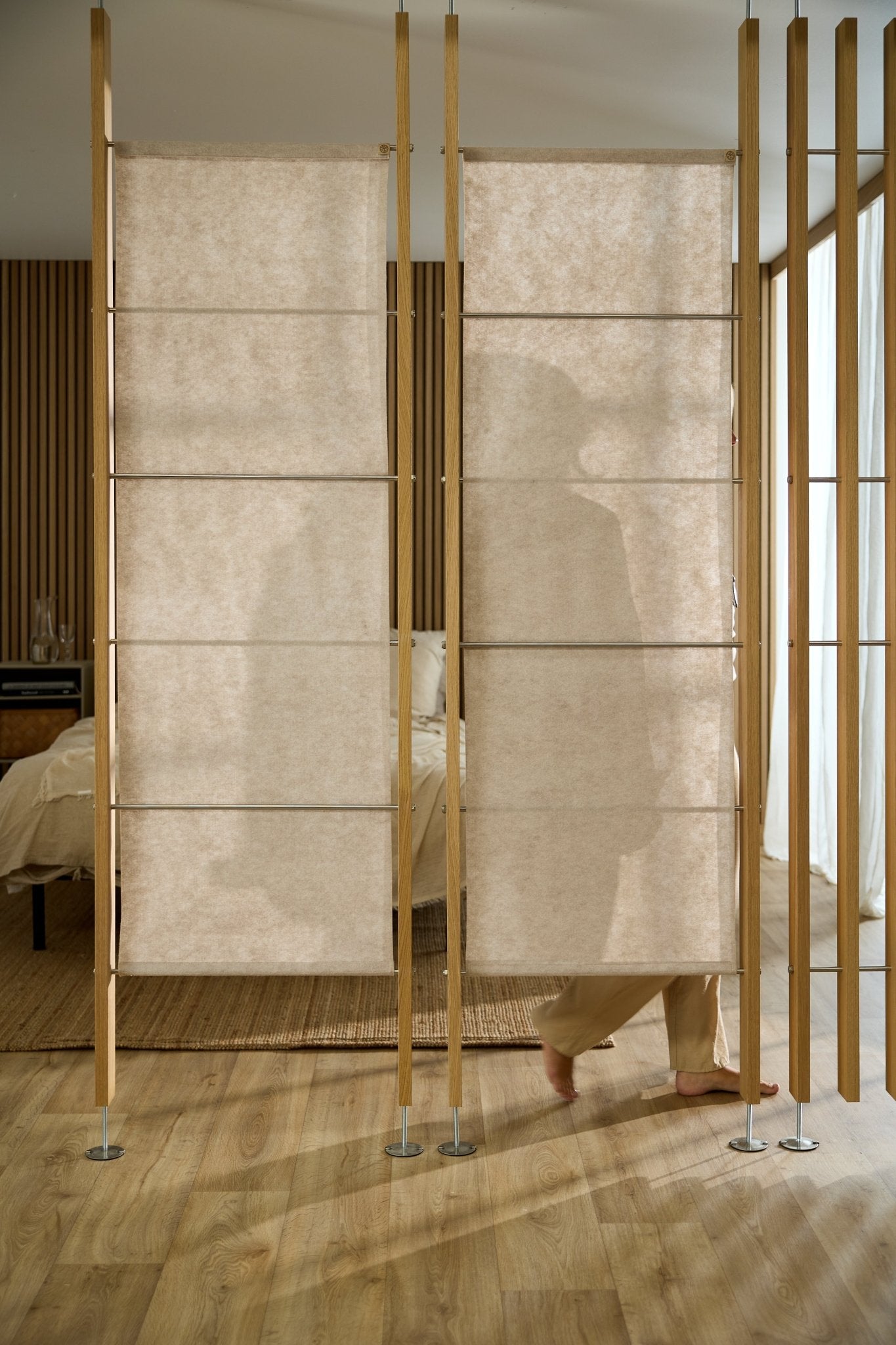 Eine Person steht hinter einer WoodUpp Vigg Felt-Akustikplatte (58 x 176 cm, Hellbeige) in einem Schlafzimmer im nordischen Stil mit Holzböden und -wänden.
