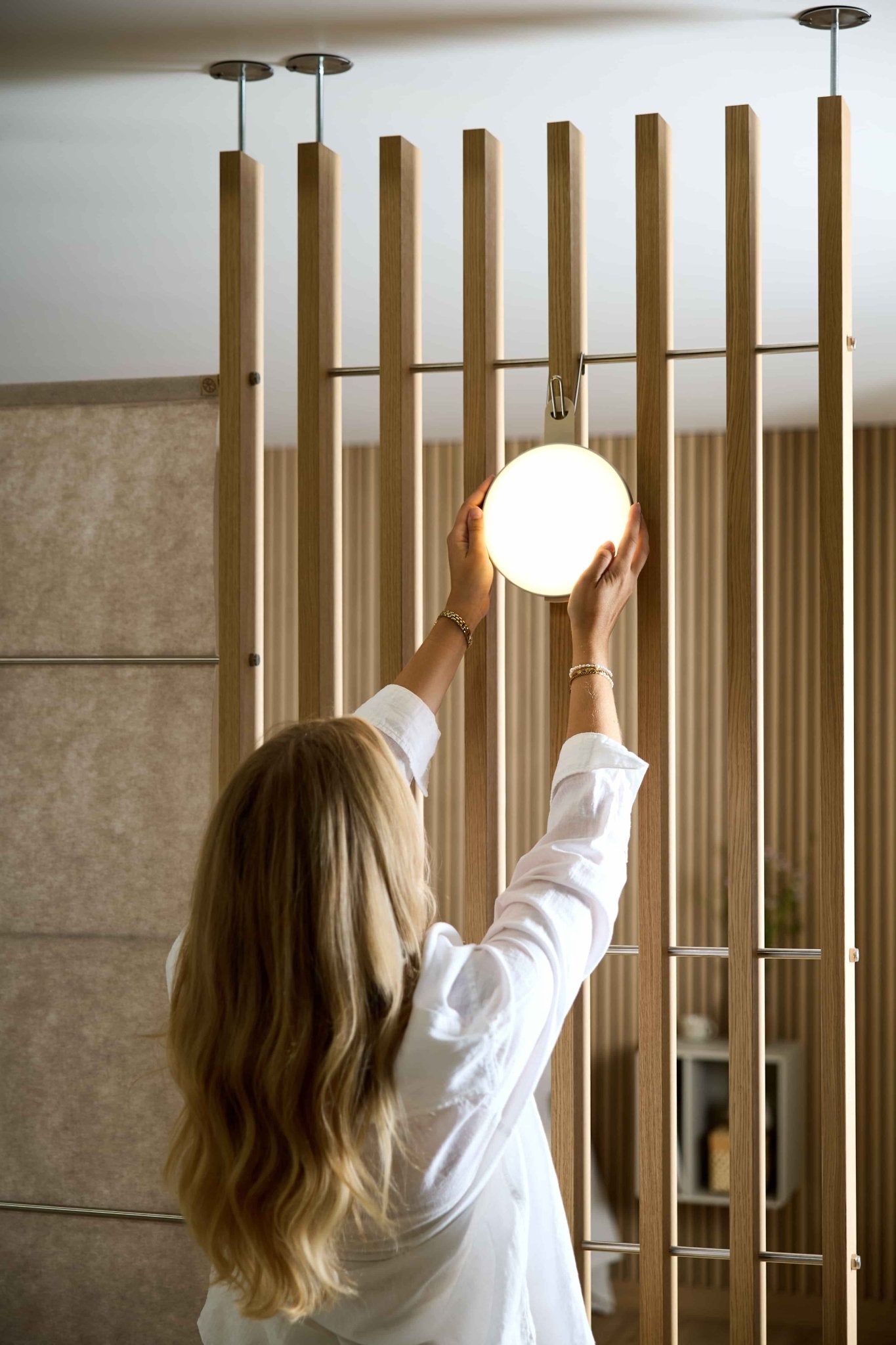 Eine Frau mit langen blonden Haaren richtet die tragbare LED-Lampe „Vala“ von WoodUpp an einem Raumteiler aus Holzlamellen der Serie „EMBRACE“ in einem modernen Interieur aus.