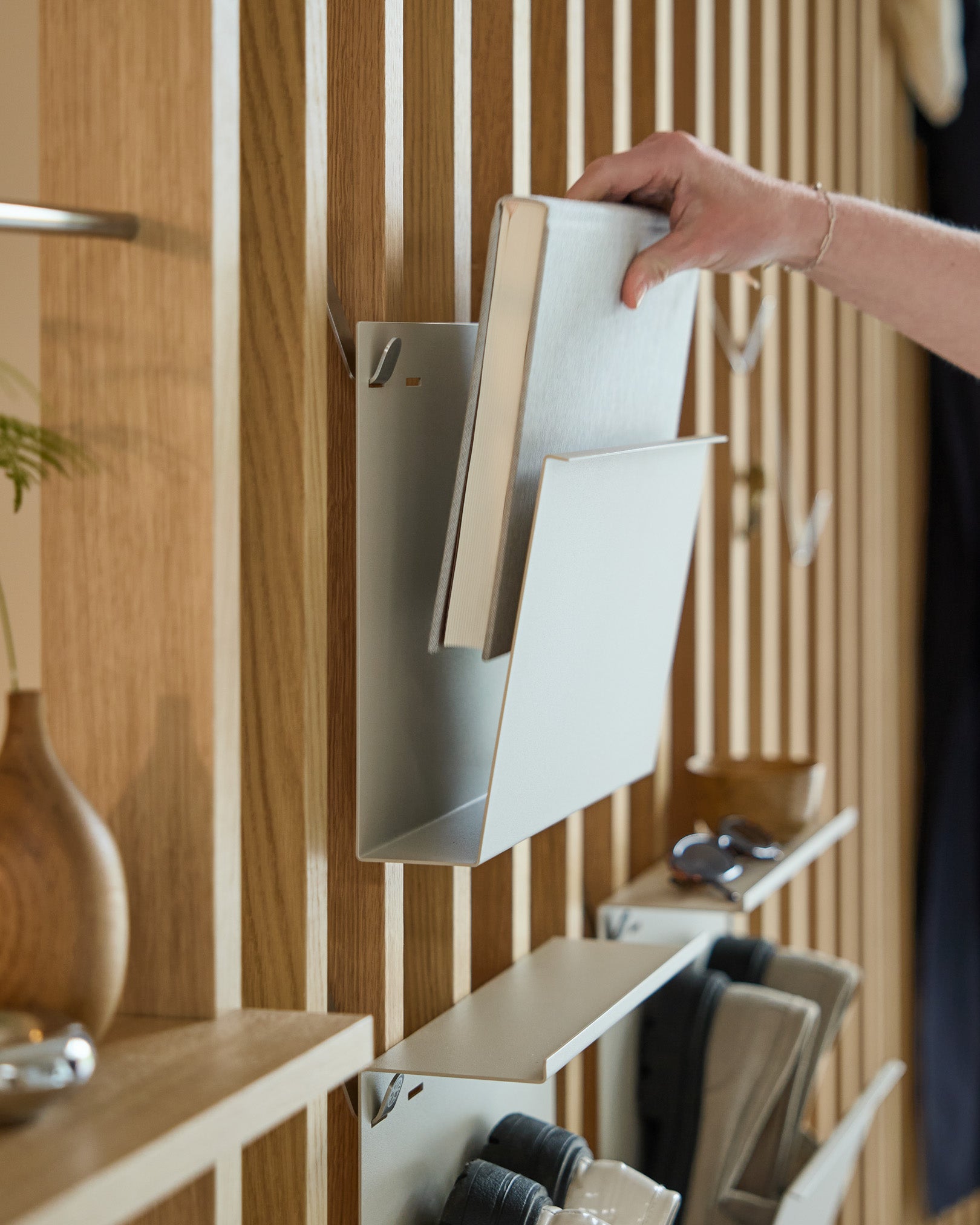 Une main place Nell | Magazine Storage / Beige de WoodUpp sur une étagère en steel thermolaqué montée sur un Panneau acoustique nordique en bois, avec des objets à proximité.