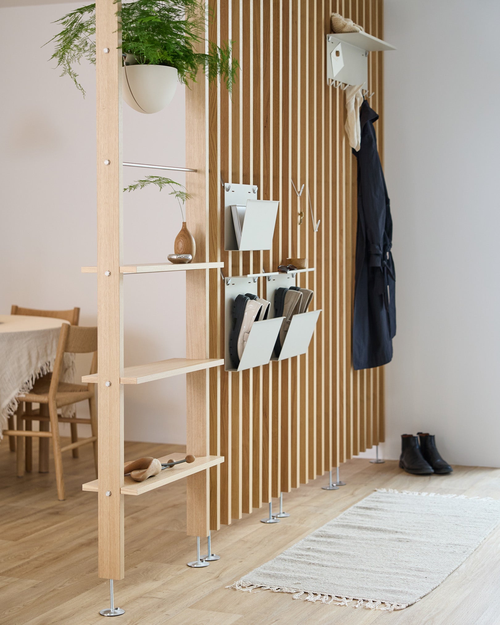 Le porte-revues Nell de WoodUpp en beige intégré à un séparateur de pièce avec des étagères en steel, des bacs de rangement et des patères ; on aperçoit derrière un coin repas et un tapis.