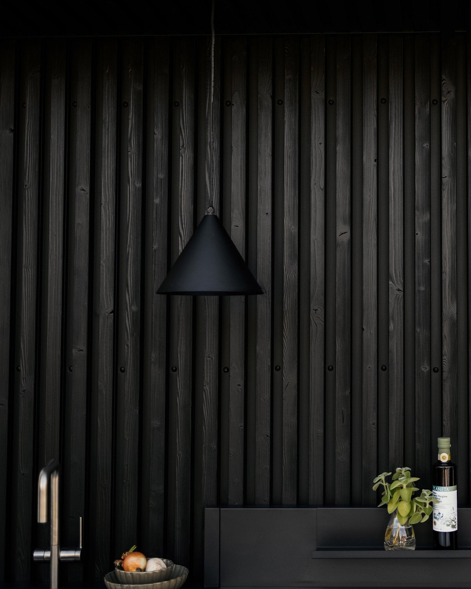 Cuisine moderne avec suspension mate black, mur acoustique en bois black texturé Akupanel, et plan de travail avec des légumes, du basilic frais et de l’huile d’olive.