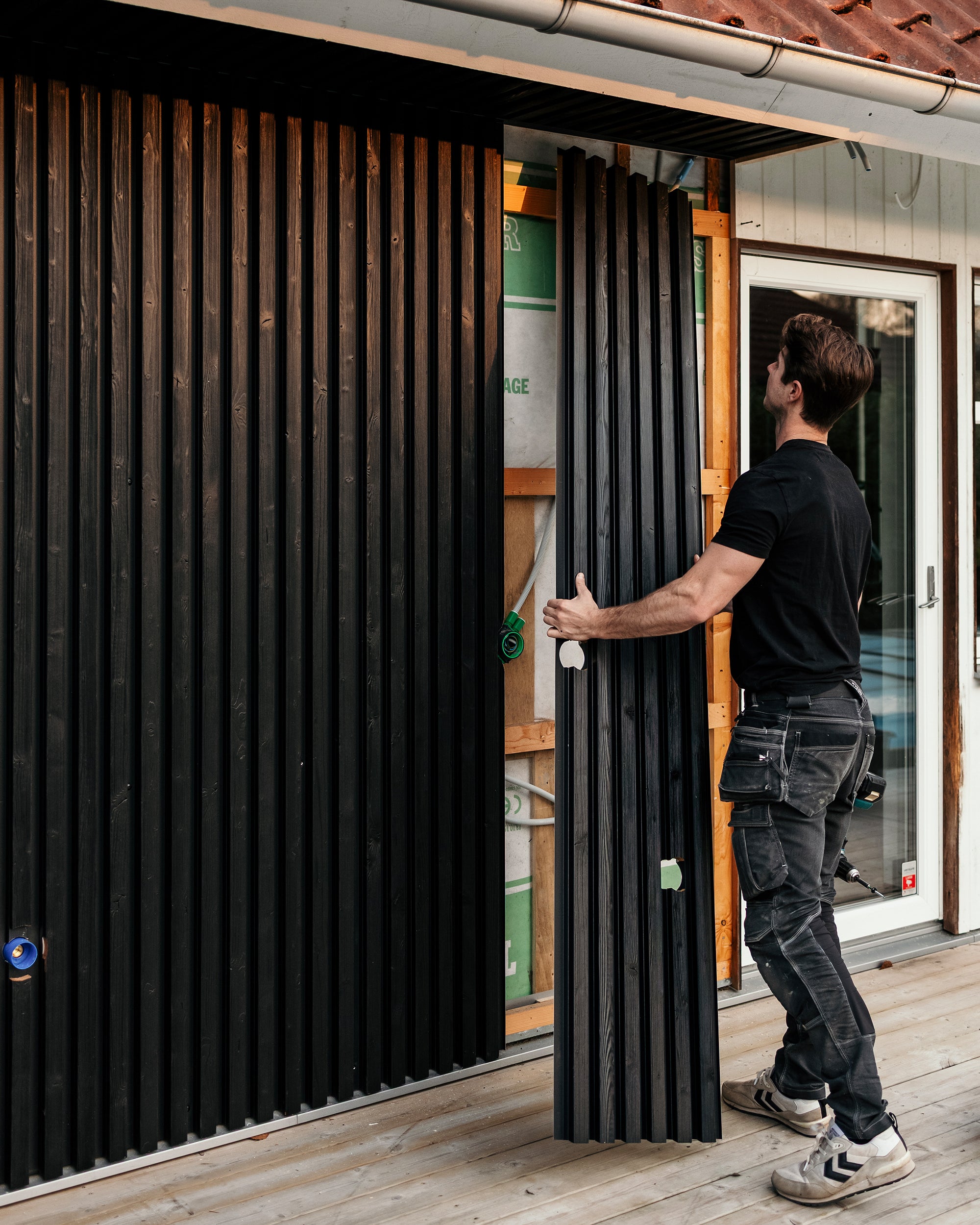 Un homme installe des panneaux de façade Aluwood noirs en position verticale sur le mur extérieur d'une maison pendant qu'il se tient sur une terrasse récemment construite.