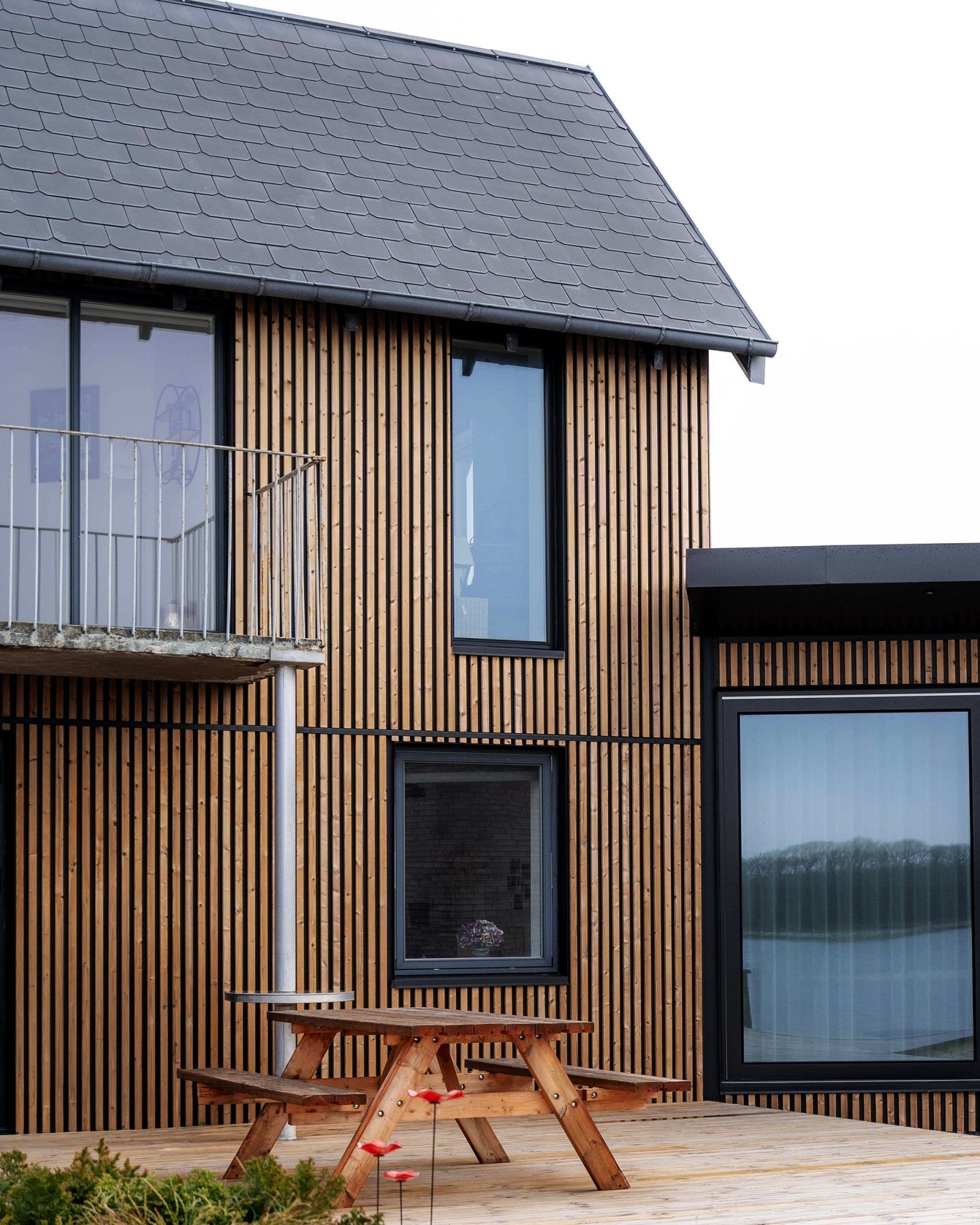Modernes Haus mit WoodUpp Aluwood Fassadenpaneelen in geöltem Brown, mit large Fenstern, einem Balkon und einer Terrasse mit Holztischbank.