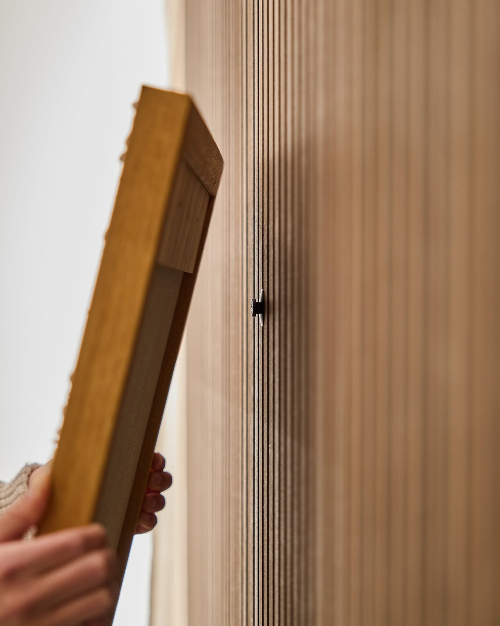 Eine Person verwendet das WoodUpp Dupps | Original-Set, um einen Rahmen für ein Akupanel Akustik-Holzpaneel zu montieren, wobei der Fokus auf dem Anbringen eines small black Hakens nahe verticalen Lamellen liegt.