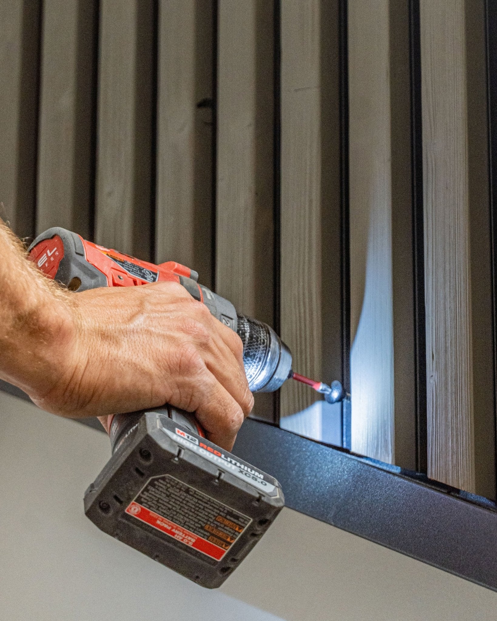 Une personne fixe une vis frontale WoodUpp black (4.8x35, rondelle Ø9.5) dans un panneau de façade Aluwood vertical à l’aide d’une perceuse-visseuse sans fil.