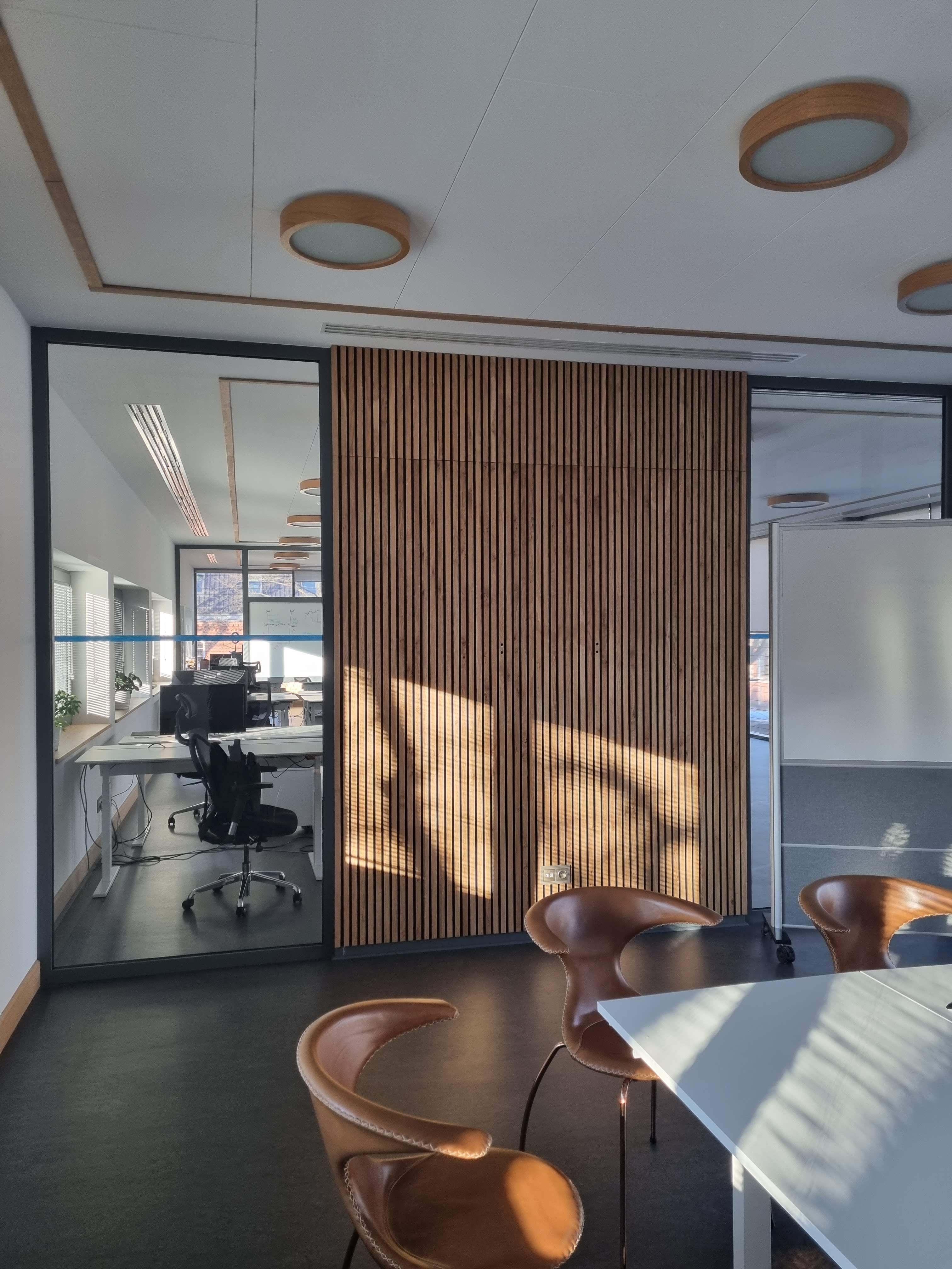 Modern office with Akupanel acoustic wood slat wall, glass partition, sunlight on brown chairs and white table, creating a warm, contemporary Nordic space.