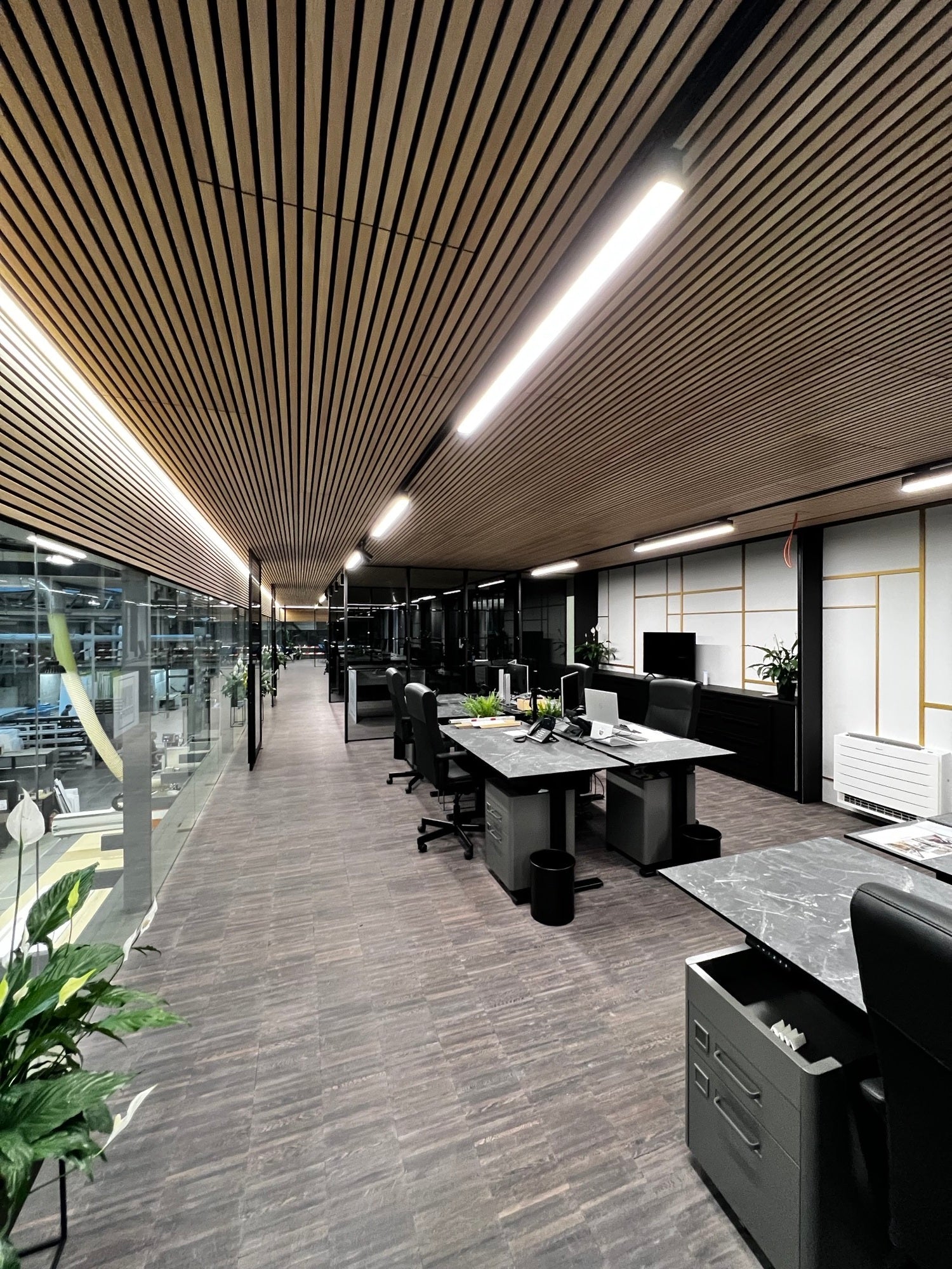 Modern office featuring Akupanel wood slat ceiling, glass wall, minimalist desks with computers, black chairs, plants, and Nordic-inspired workspace design.