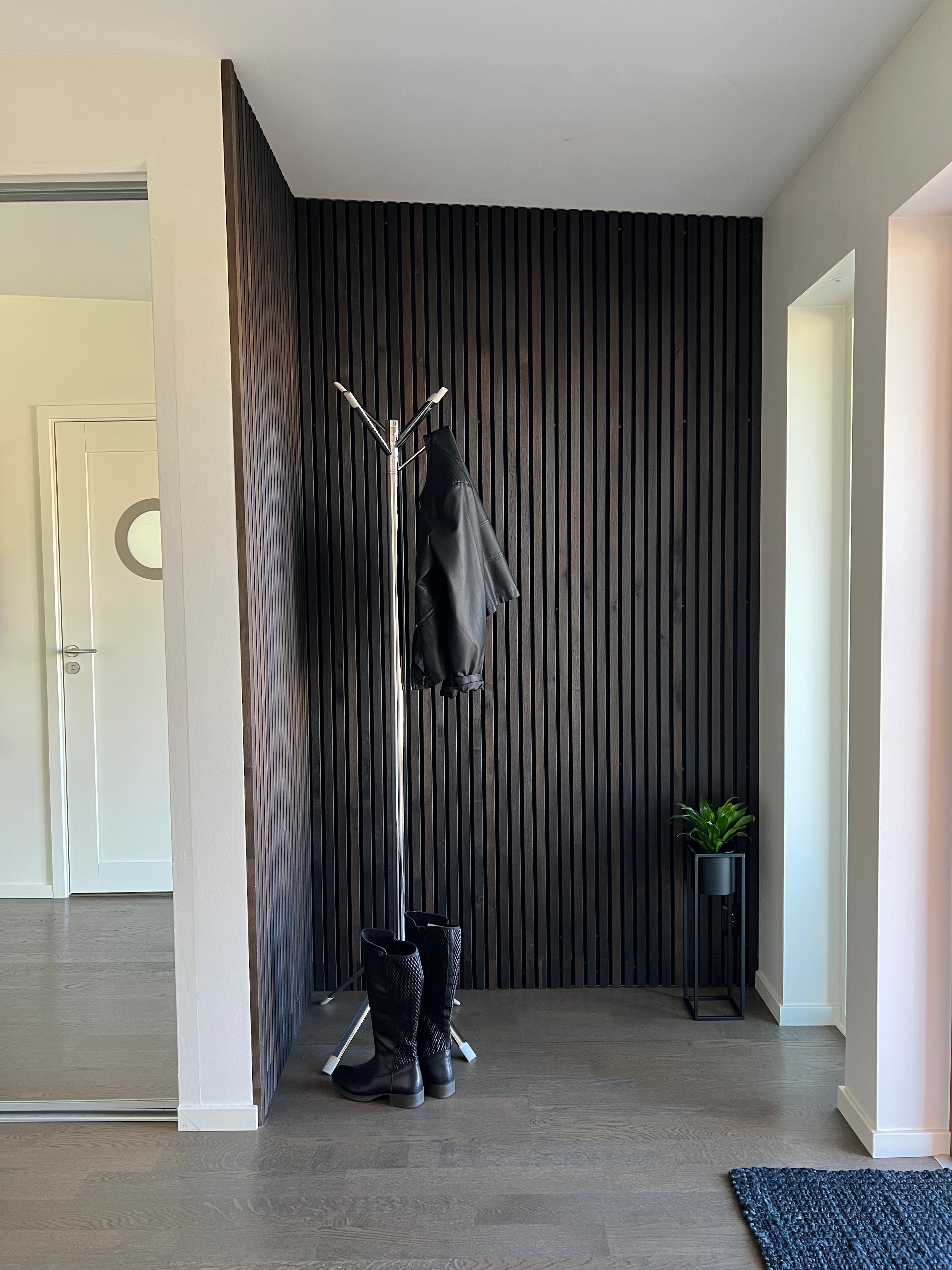 Minimalist entryway featuring a black coat rack with jacket and boots, small plant, Akupanel dark wood acoustic paneling, and light walls and floor.