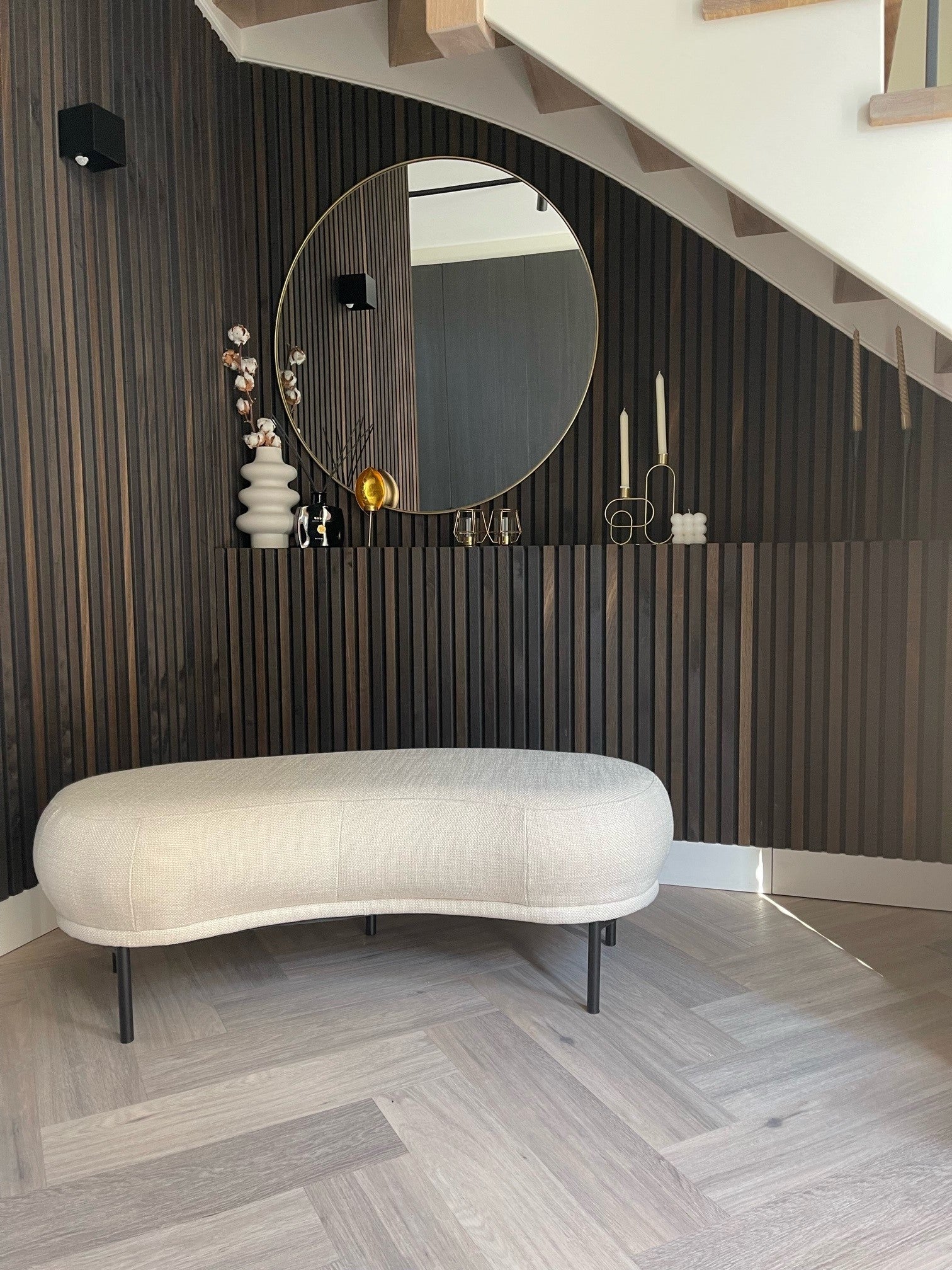 Modern entryway with a curved beige bench, round mirror, black Akupanel acoustic wood slat wall, and decorative vases and candles under the staircase.