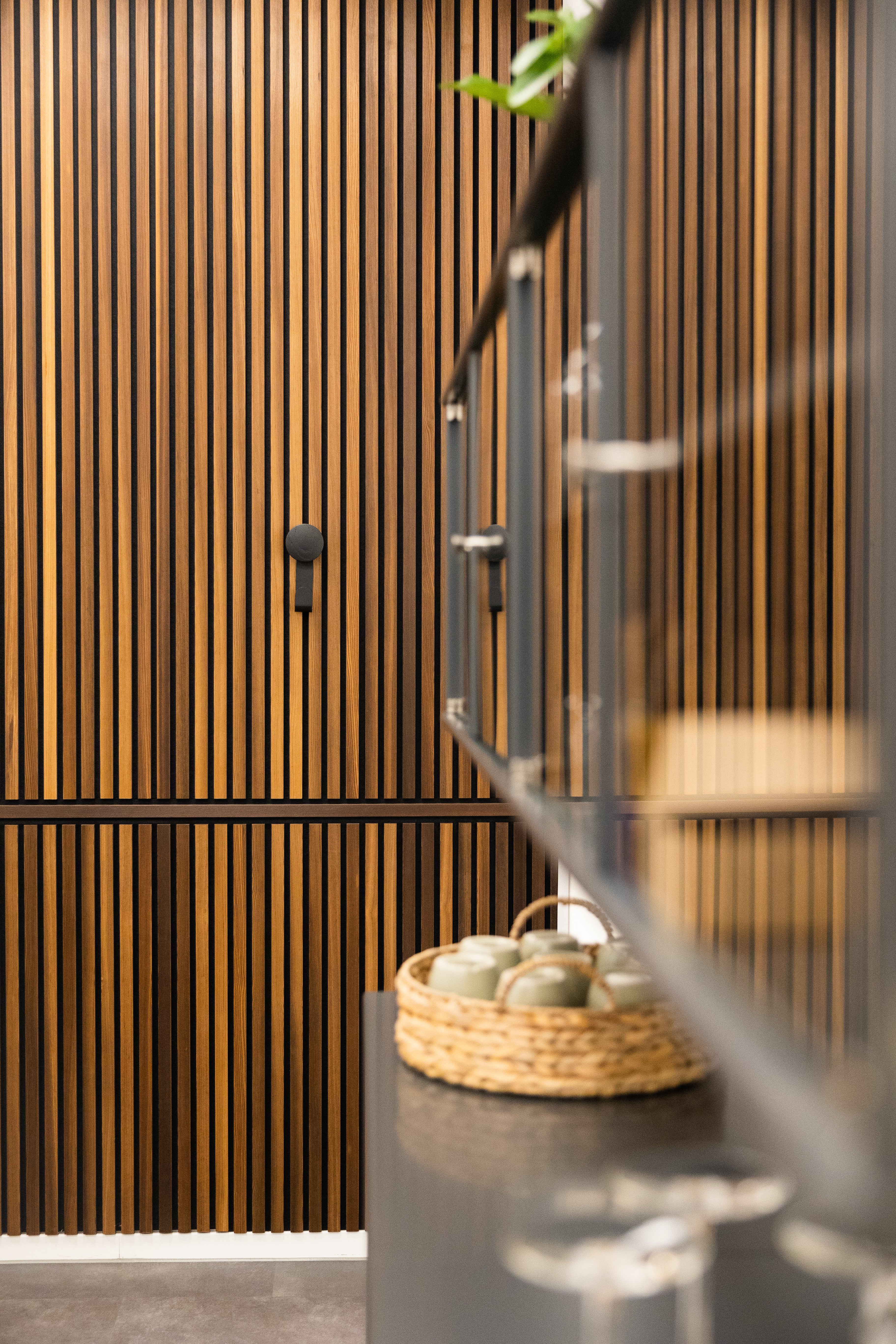 Modern kitchen with a vertical acoustic slat wall panel, black countertop, glass cabinet, and wicker basket holding mugs for a Nordic-inspired look.