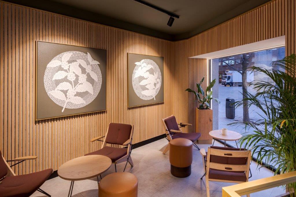Modern lounge with Nordic acoustic wood panels, abstract leaf art, brown chairs, round tables, and a large window overlooking street and lush plants.