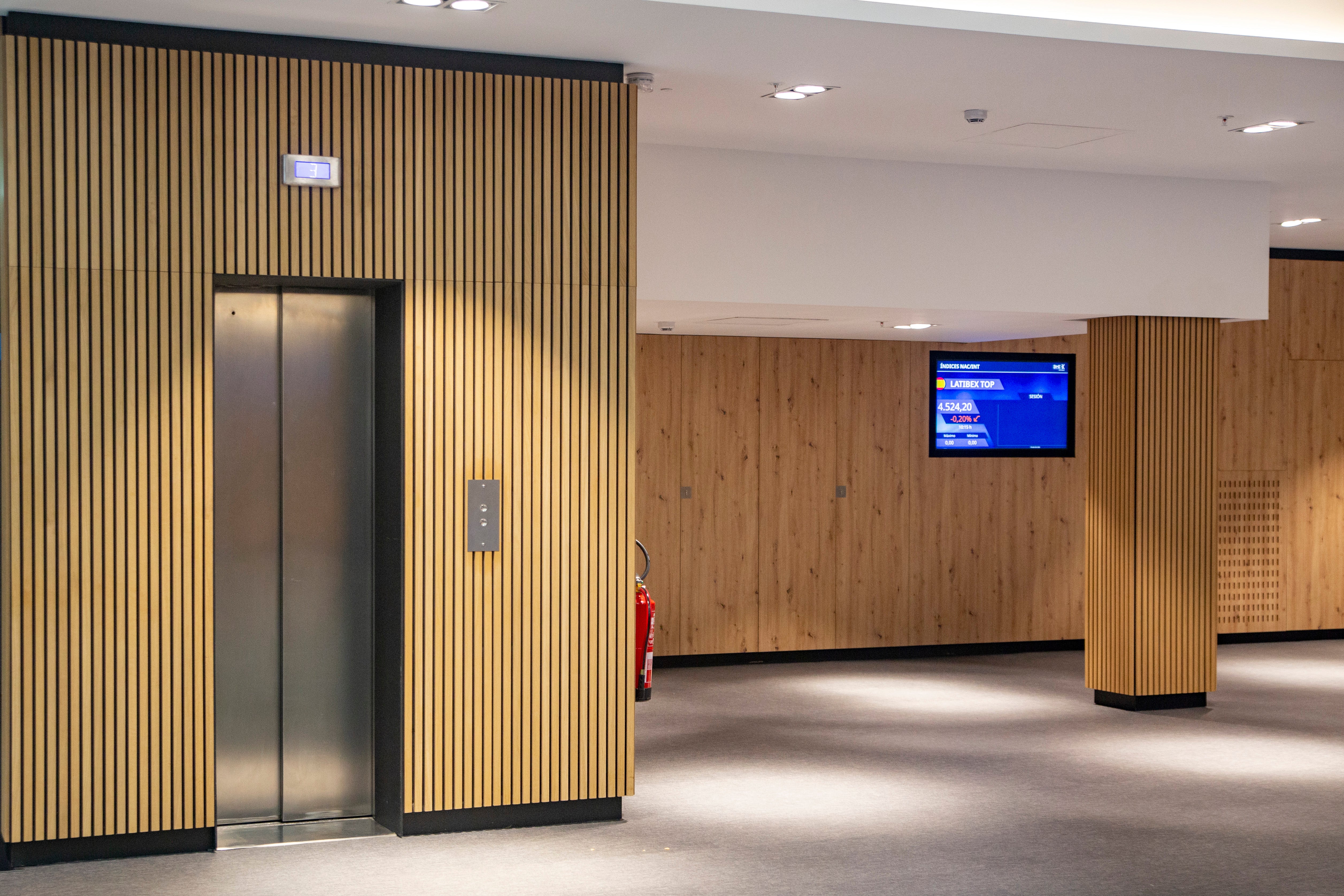 Modern hallway with closed elevator door, Akupanel acoustic wood paneling by WoodUpp on walls, fire extinguisher, and digital TV information screen.