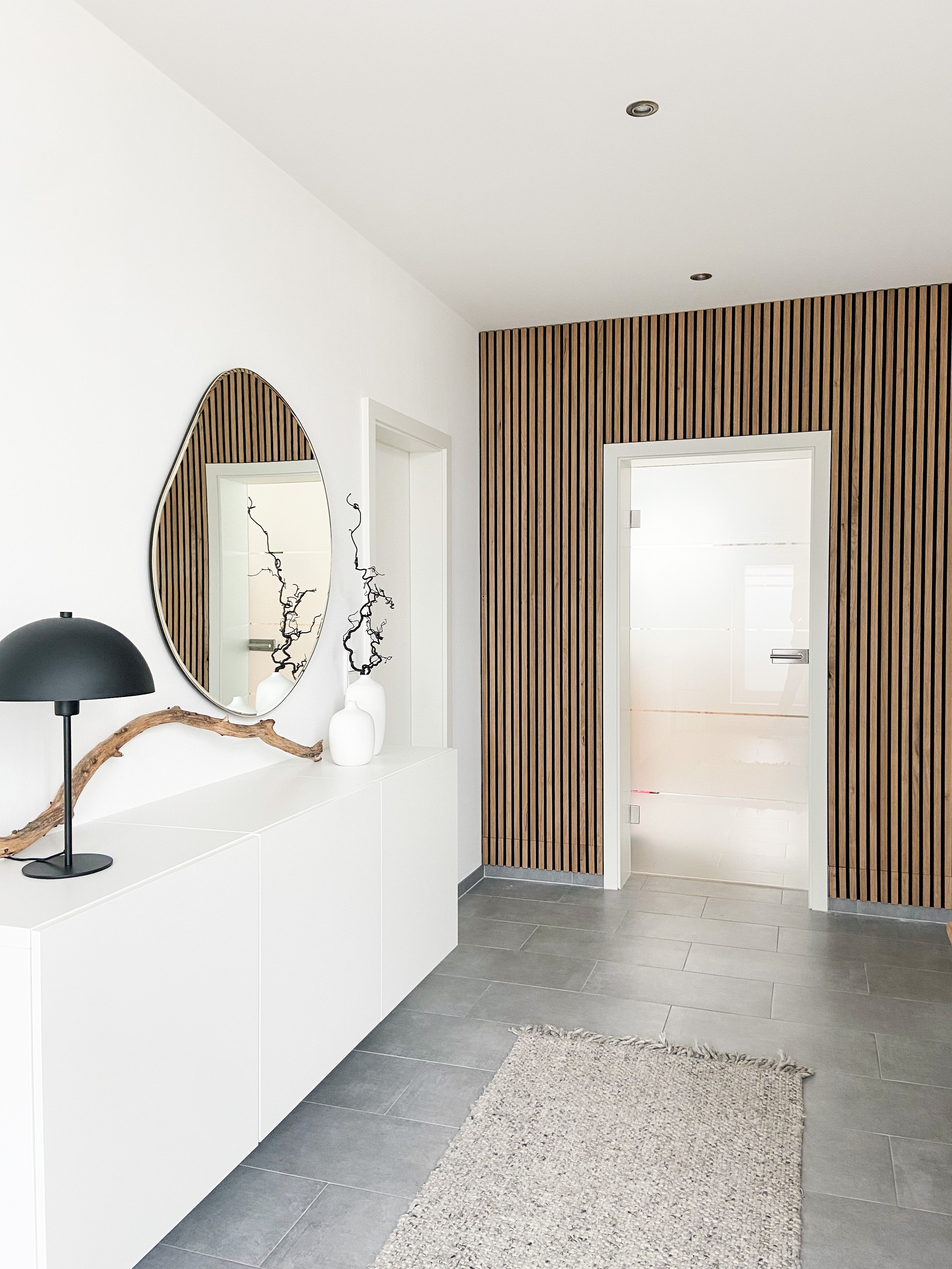 Minimalist hallway with Akupanel acoustic wood slat accent wall, white cabinet, black lamp, decorative branch, oval mirror, gray tiled floor, and textured rug.