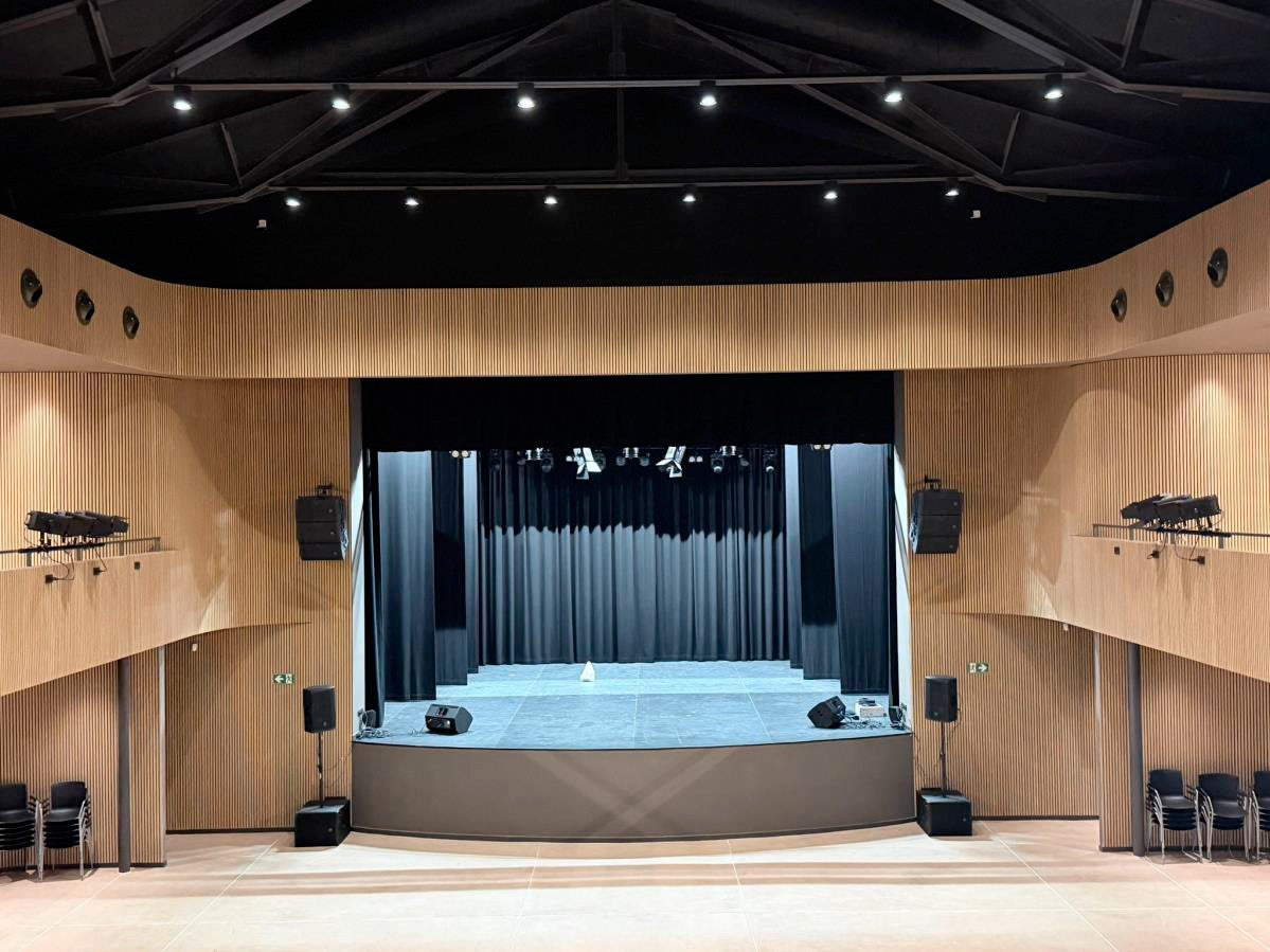Modern theater with Nordic wooden acoustic panels, empty stage, black curtains, chairs stacked at sides, and overhead lighting illuminating the interior.
